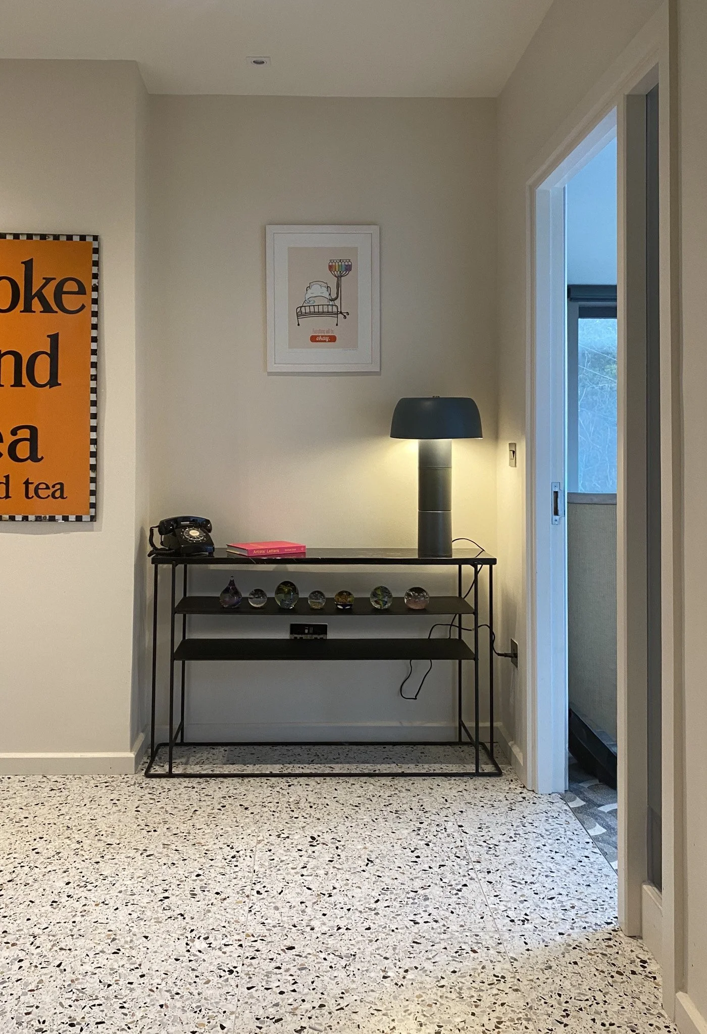 A minimalist black metal console table with glass decor items, a black table lamp, an old-fashioned black rotary telephone, a pink book, and a black power strip on it, set against a white wall with framed art and a larger colorful poster, and a water