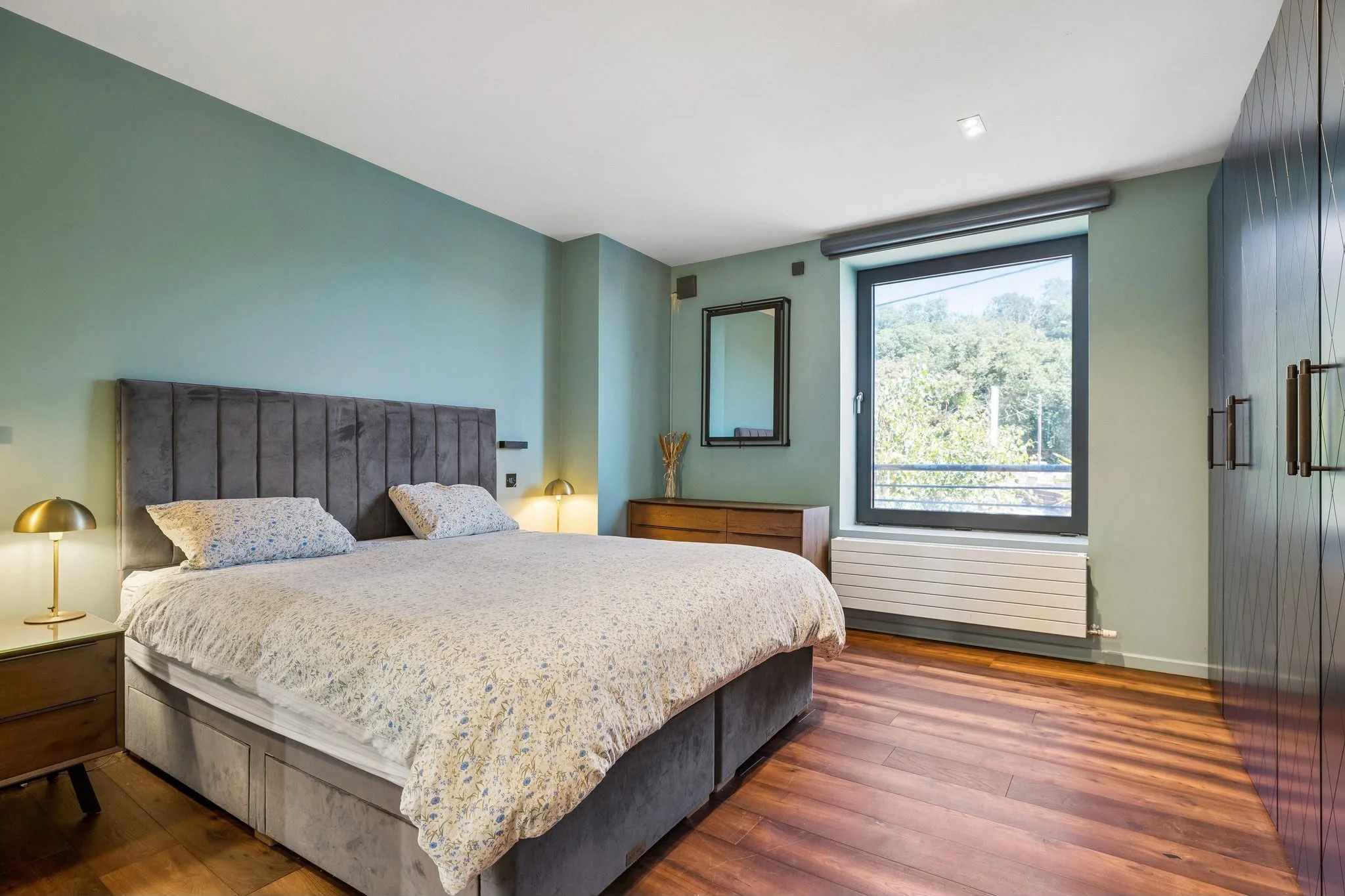 A bedroom with a large bed, a headboard, nightstands, a lamp, and a window showing greenery outside.