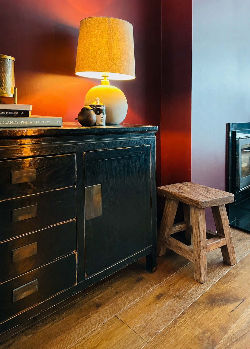 A corner of a room with a black wooden cabinet, a table lamp with a beige shade, a small decorative object, and a stack of books. There's a rustic wooden stool and a part of a glass-fronted appliance. The wall behind is painted dark red.