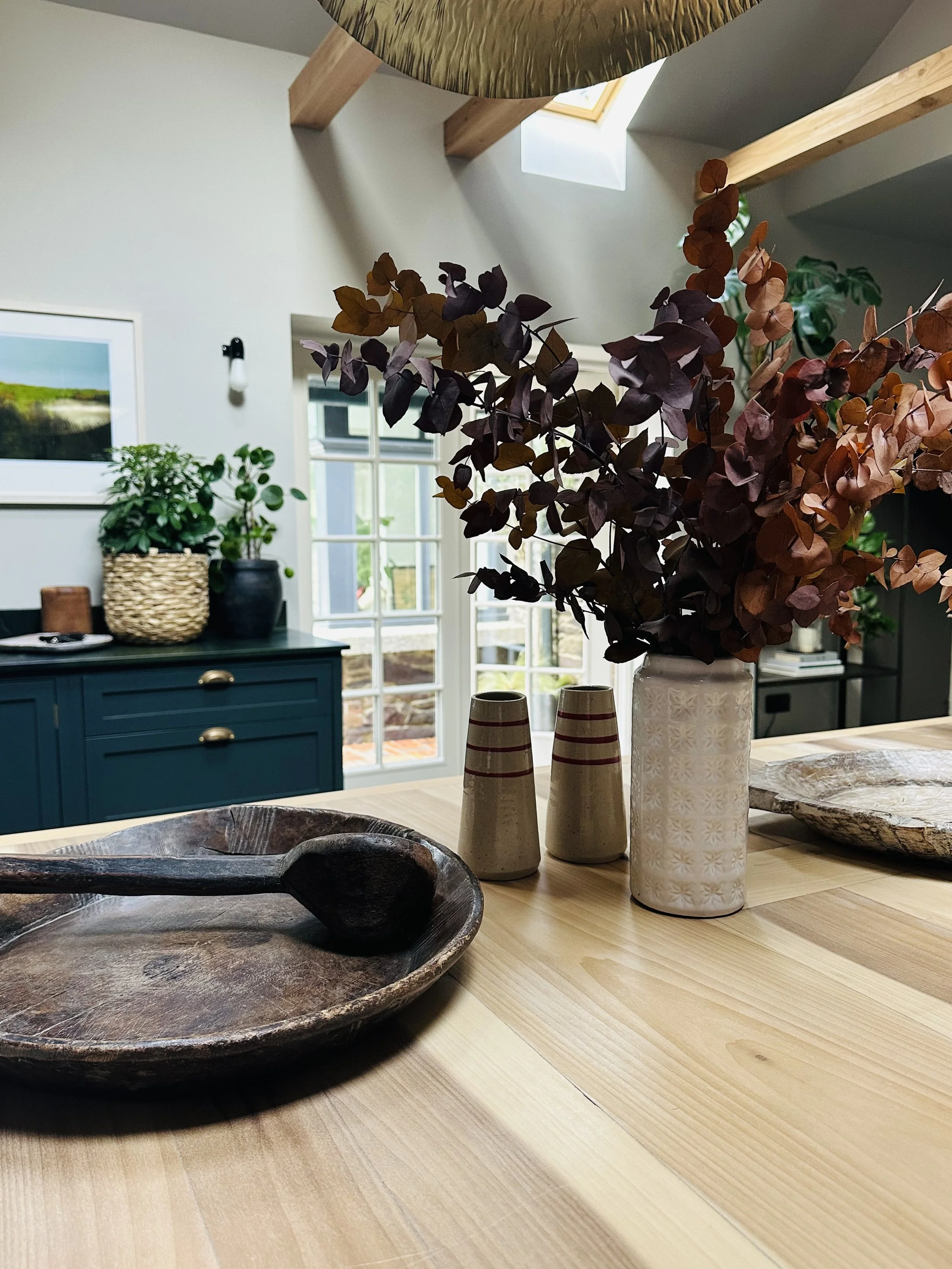 A wooden table with decorative vases and bowls in a bright room with a window, plants, and artwork on the walls.