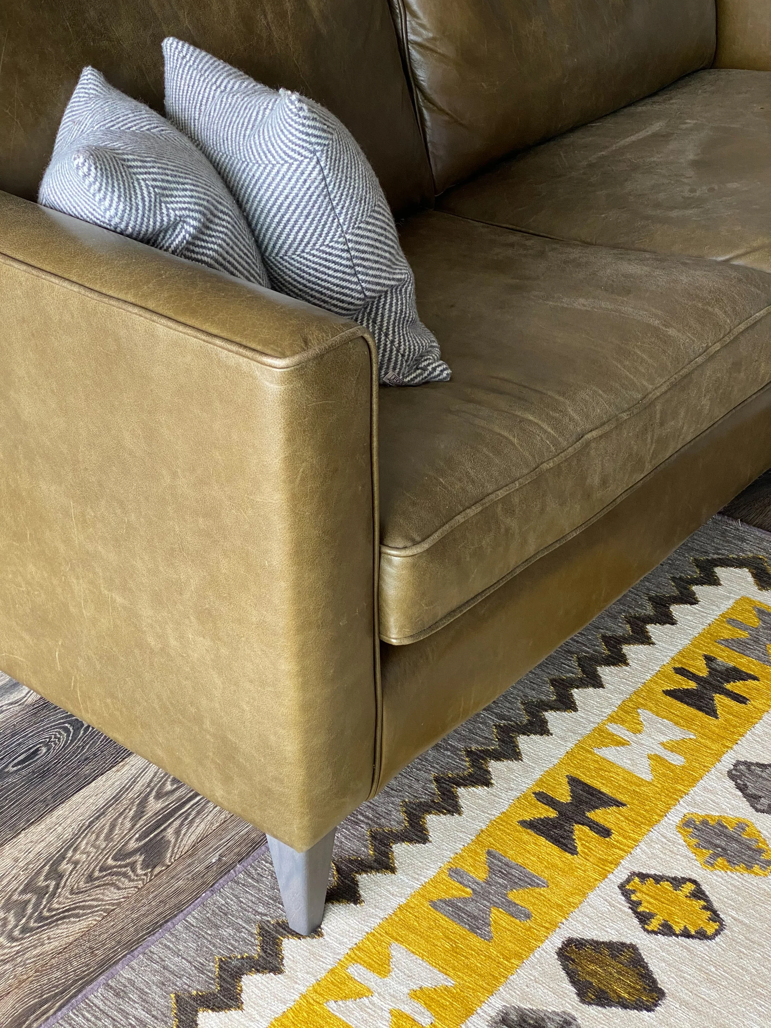 Close-up of a brown leather couch with two blue and white checked throw pillows, a yellow and gray patterned rug on a dark wood floor.