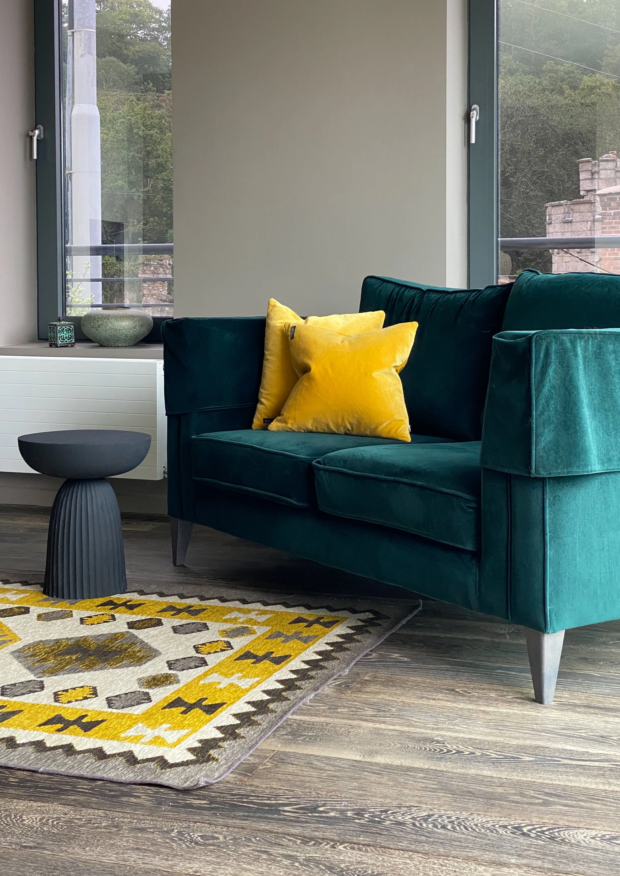 Living room with a green velvet sofa, yellow pillows, a black side table, a colorful patterned rug, and large windows showing trees and a brick building outside.
