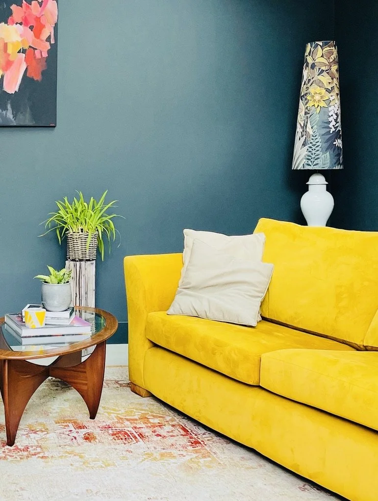 Bright yellow sofa with a white pillow, next to a wooden coffee table with plants and books, against a teal wall with a floral lamp and colorful artwork.
