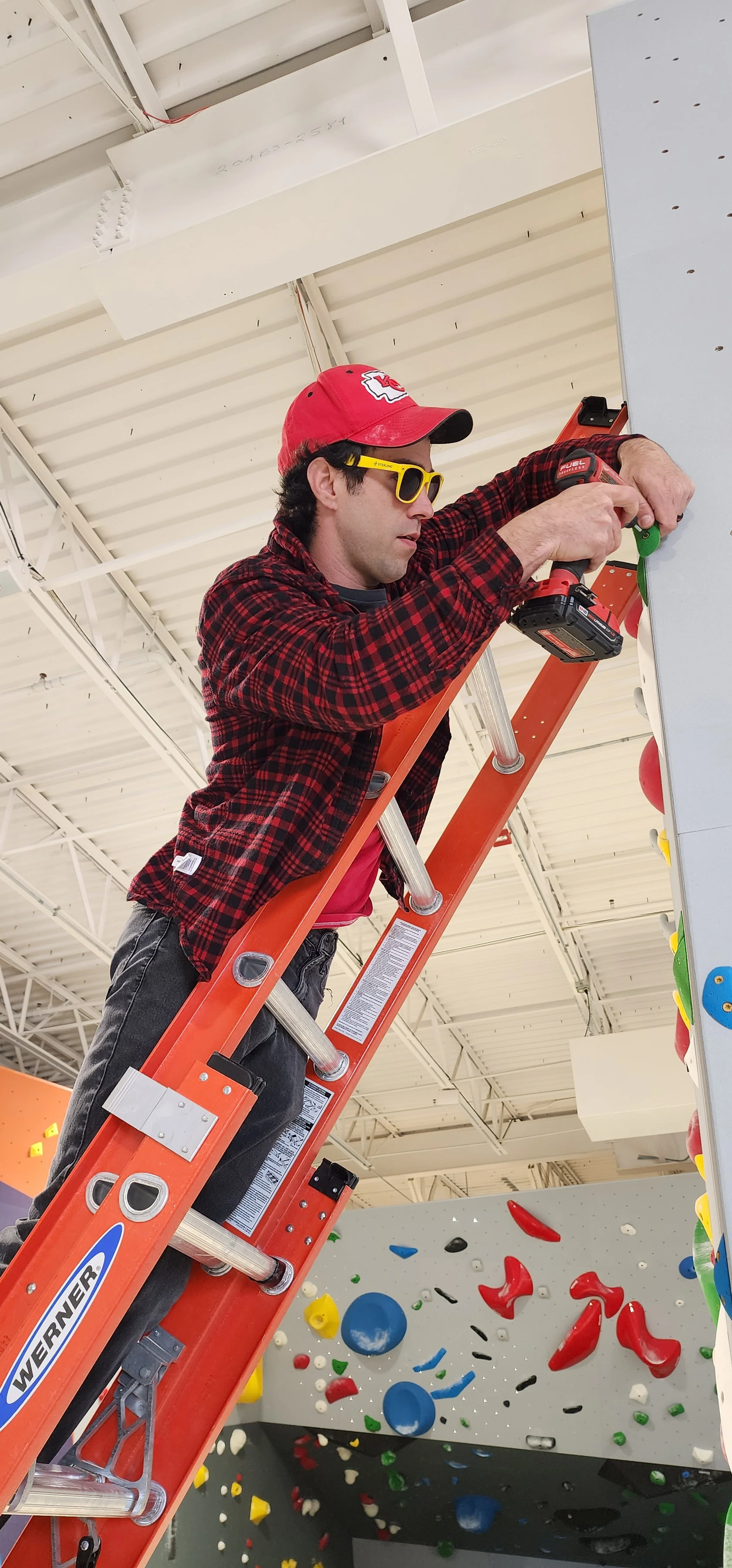 Person on an orange ladder using a power drill to install climbing holds on an indoor climbing wall, wearing a red plaid shirt and red cap.