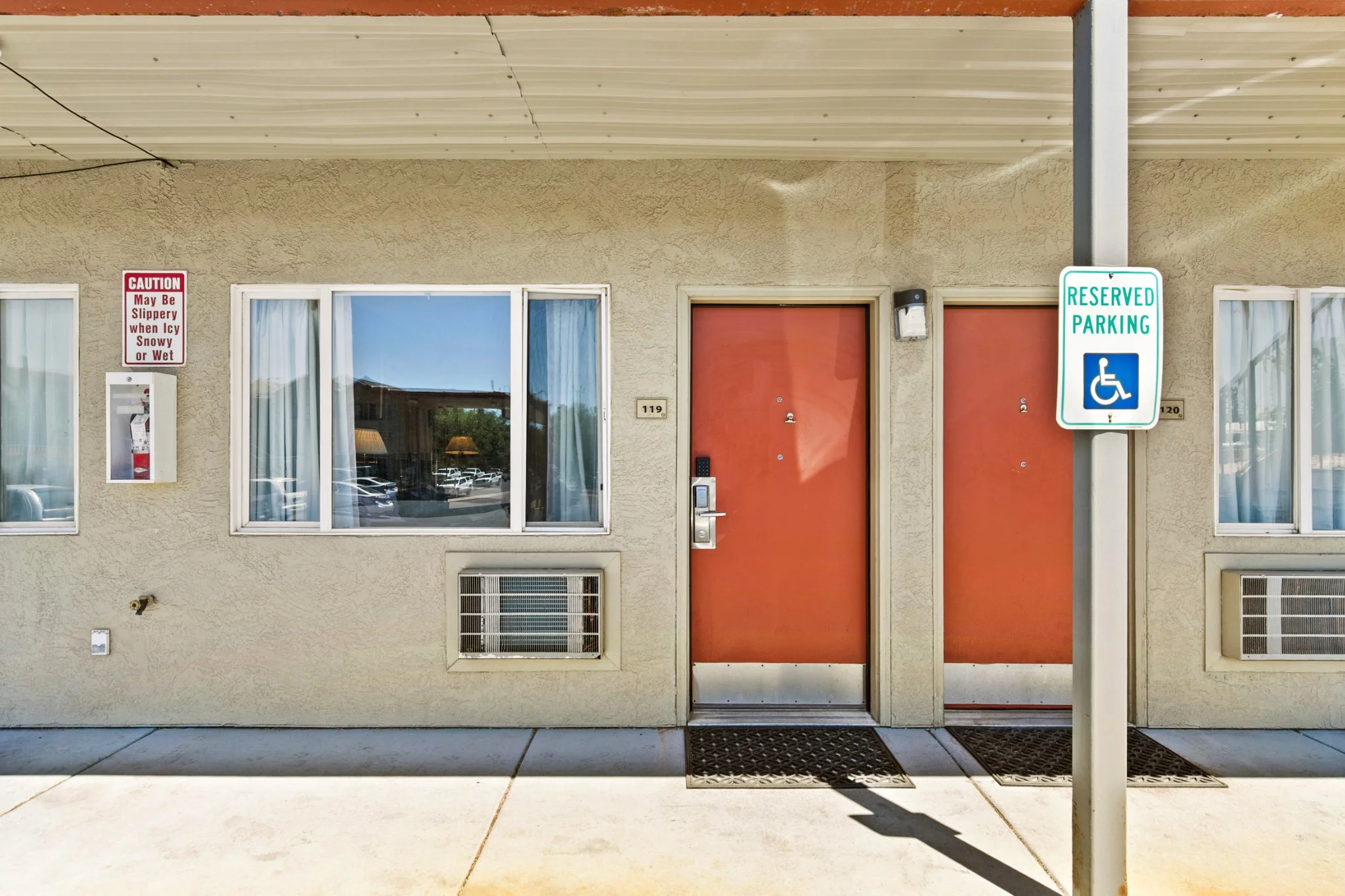 Exterior of a building with a beige stucco wall, three windows, and a red door, featuring a reserved parking sign with a wheelchair symbol. There is a caution sign on the wall indicating slipperiness when icy, snowy, or wet. The sidewalk and black ru