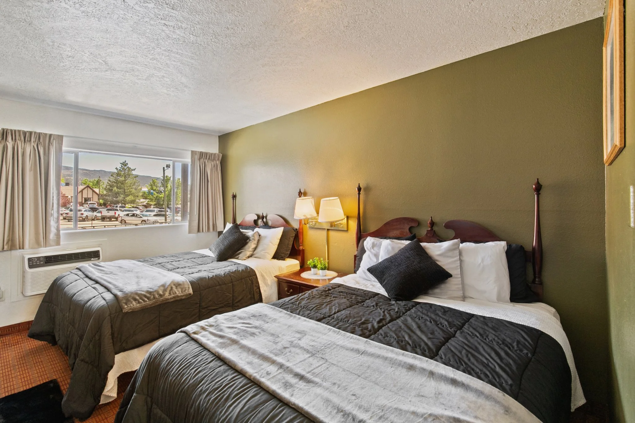 A hotel room with two double beds covered in dark gray bedspreads and pillows, a bedside table with a lamp, and a window showing a parking lot and trees outside.