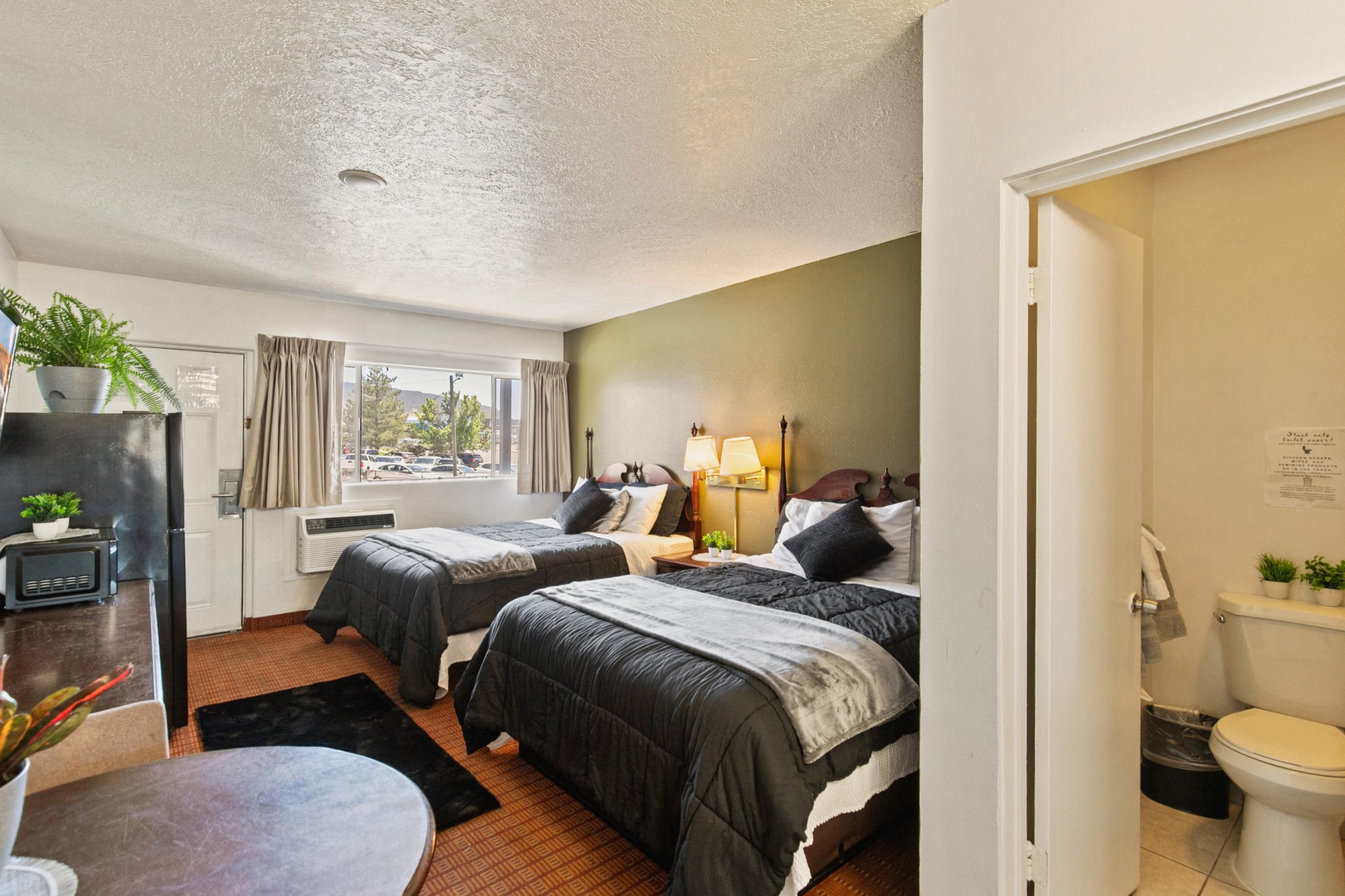 A hotel room with two queen beds, nightstands, lamps, window with curtains, and a small room with toilet and sink visible through an open door.