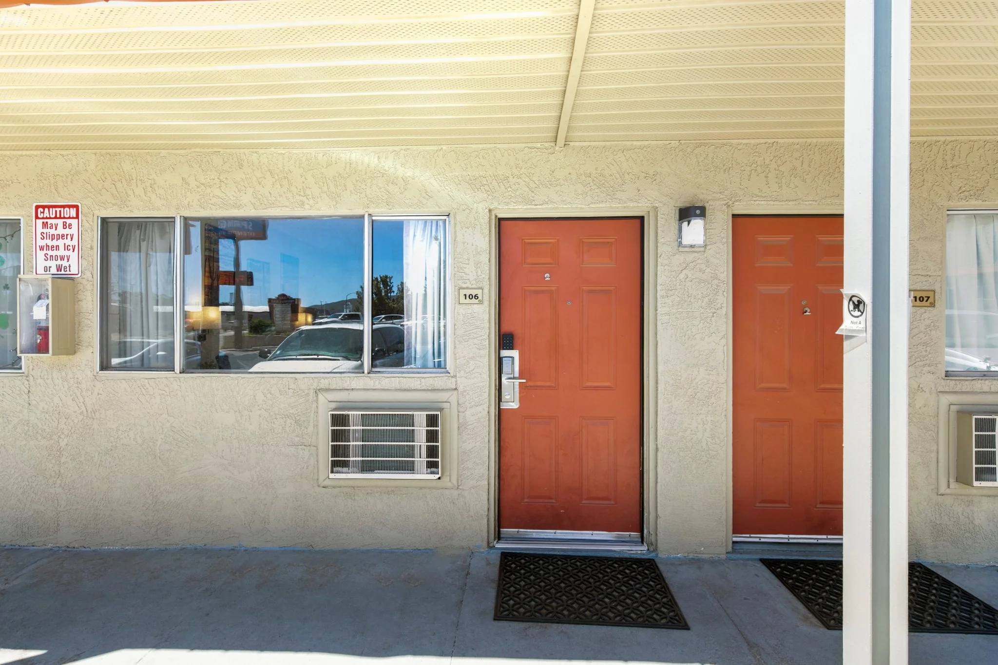 Exterior view of two adjacent apartments with orange doors numbered 106 and 107, two windows, a sign warning of slippery surfaces in winter, a mailbox, and an air vent below the window.