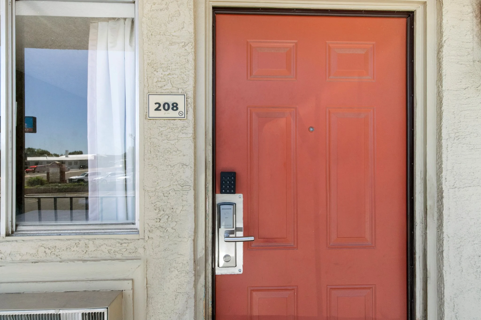 Red front door with a peephole, digital door lock, and an occasional code keypad. To the left, a window with a white curtain partly drawn back. A house number plaque with the number 208 is mounted on the textured beige wall next to the door.