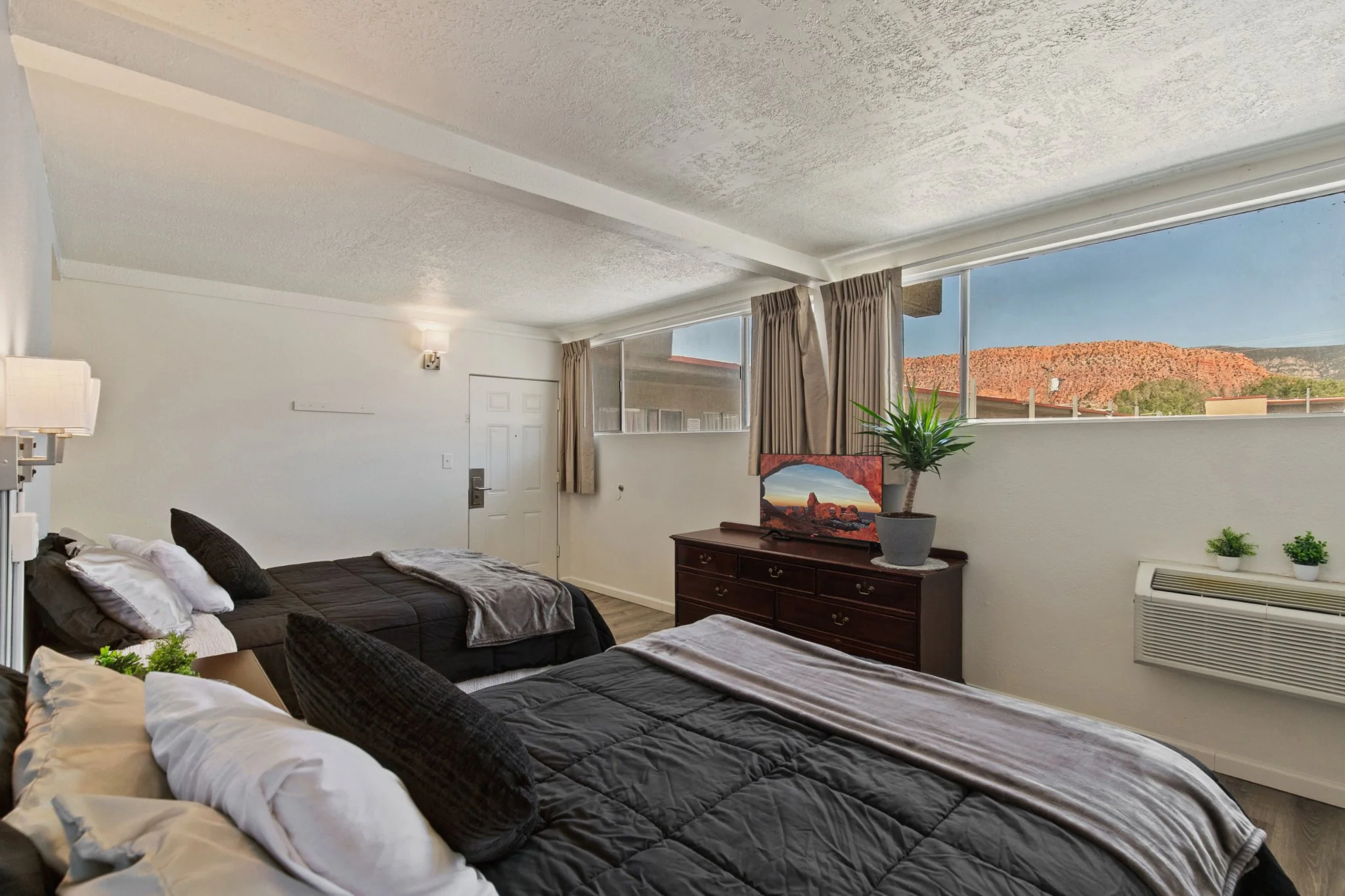 A hotel room with two beds with black and white bedding, a wooden dresser with a landscape painting, a potted plant, a lamp, and windows showing a rocky hillside and blue sky.