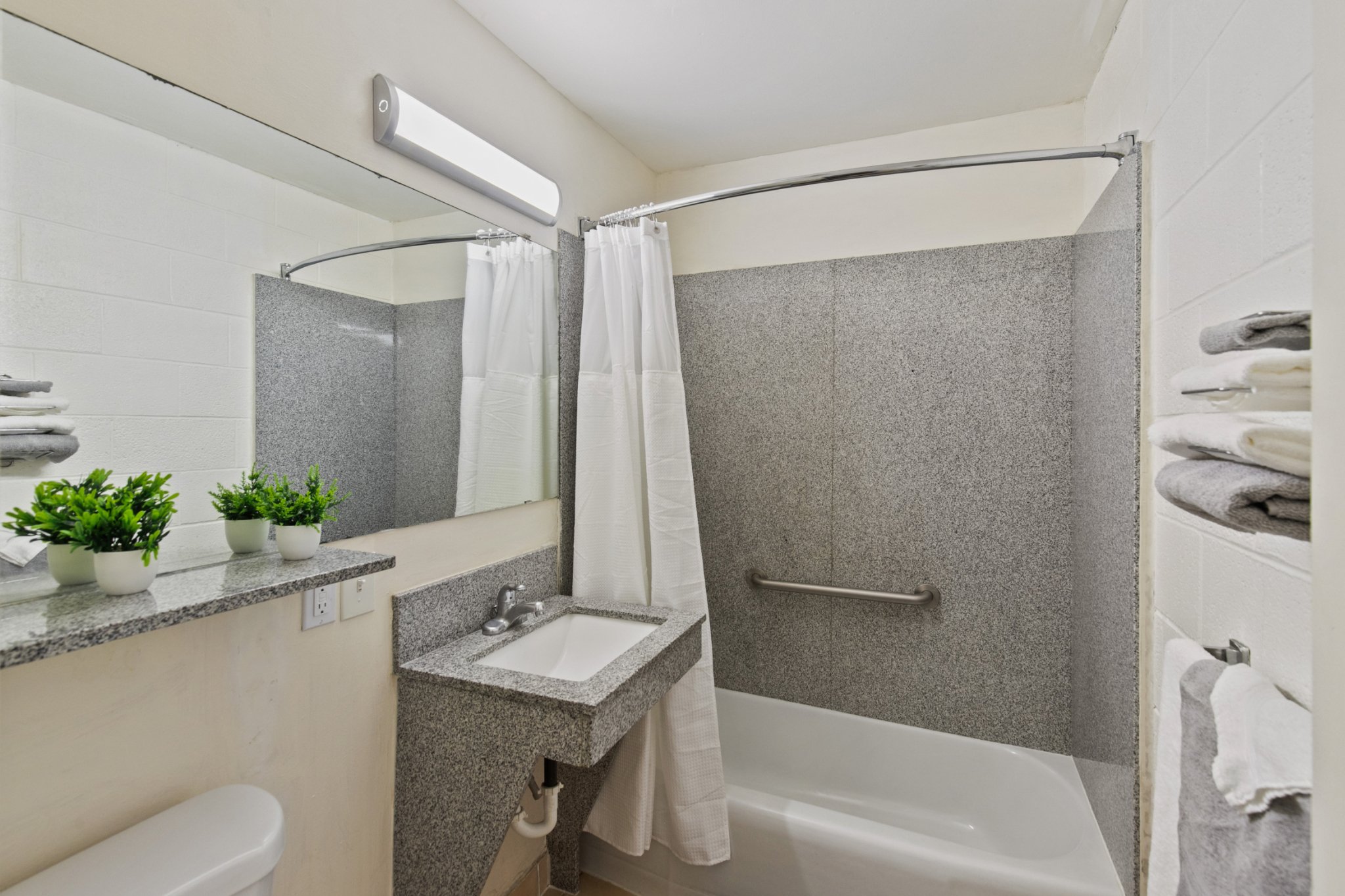 A clean bathroom with a granite countertop sink, a mirror, a floral arrangement, towel racks with folded towels, a shower with a curved curtain rod and white shower curtain, and a grab bar in the tub area.