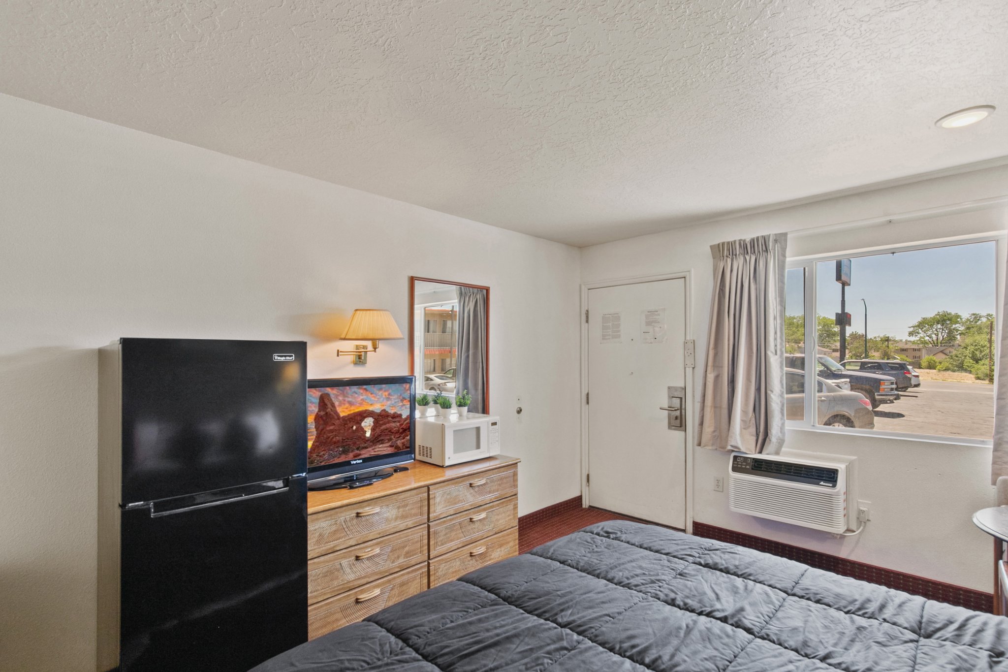 View of a motel room with a bed, wooden dresser, small TV, mini microwave, and a window with a curtain showing parked cars outside.
