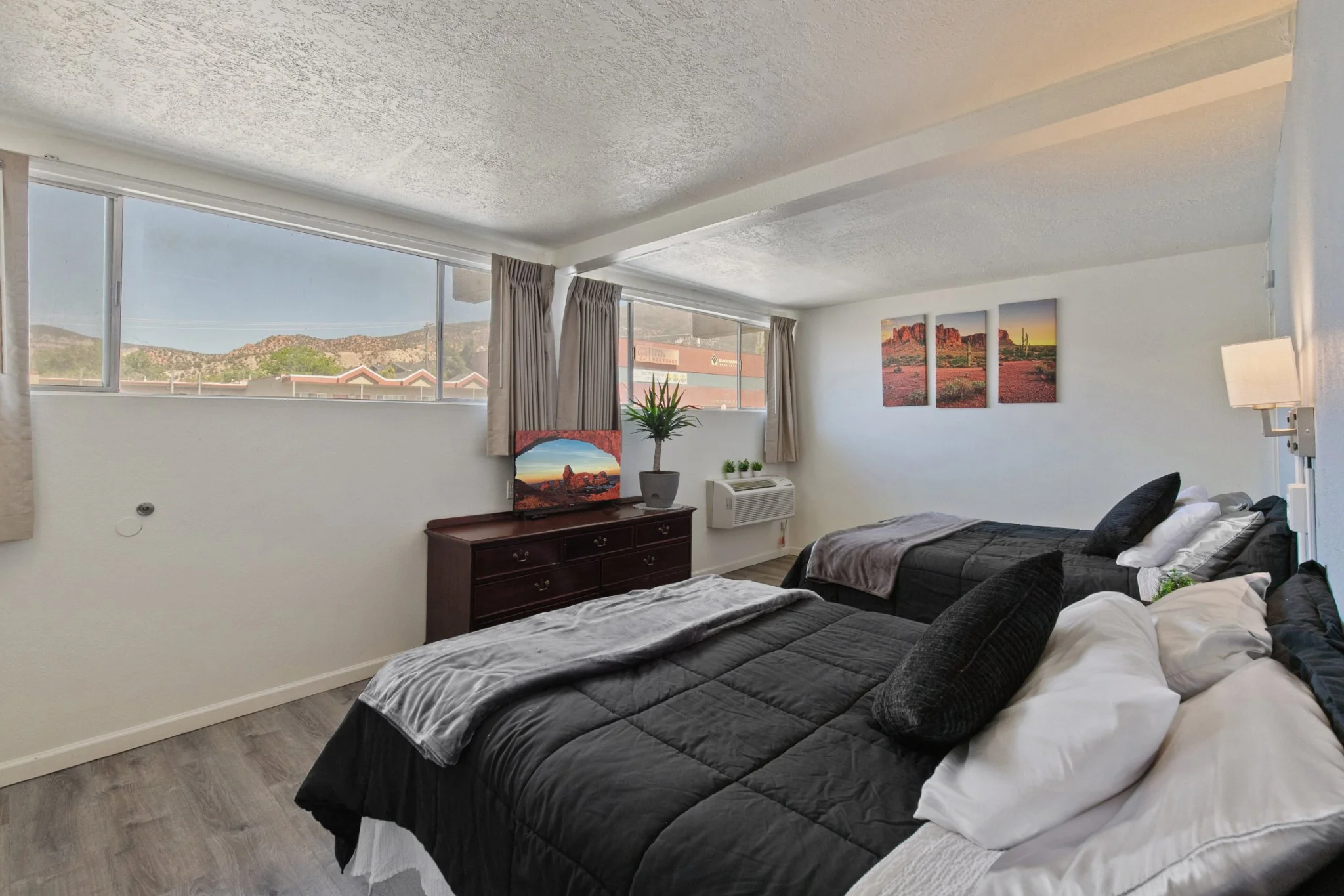 A bedroom with two beds, black bedding, and white sheets. There is a wooden dresser with paintings of desert landscapes and a potted plant on top. Large windows with curtains show mountains outside.