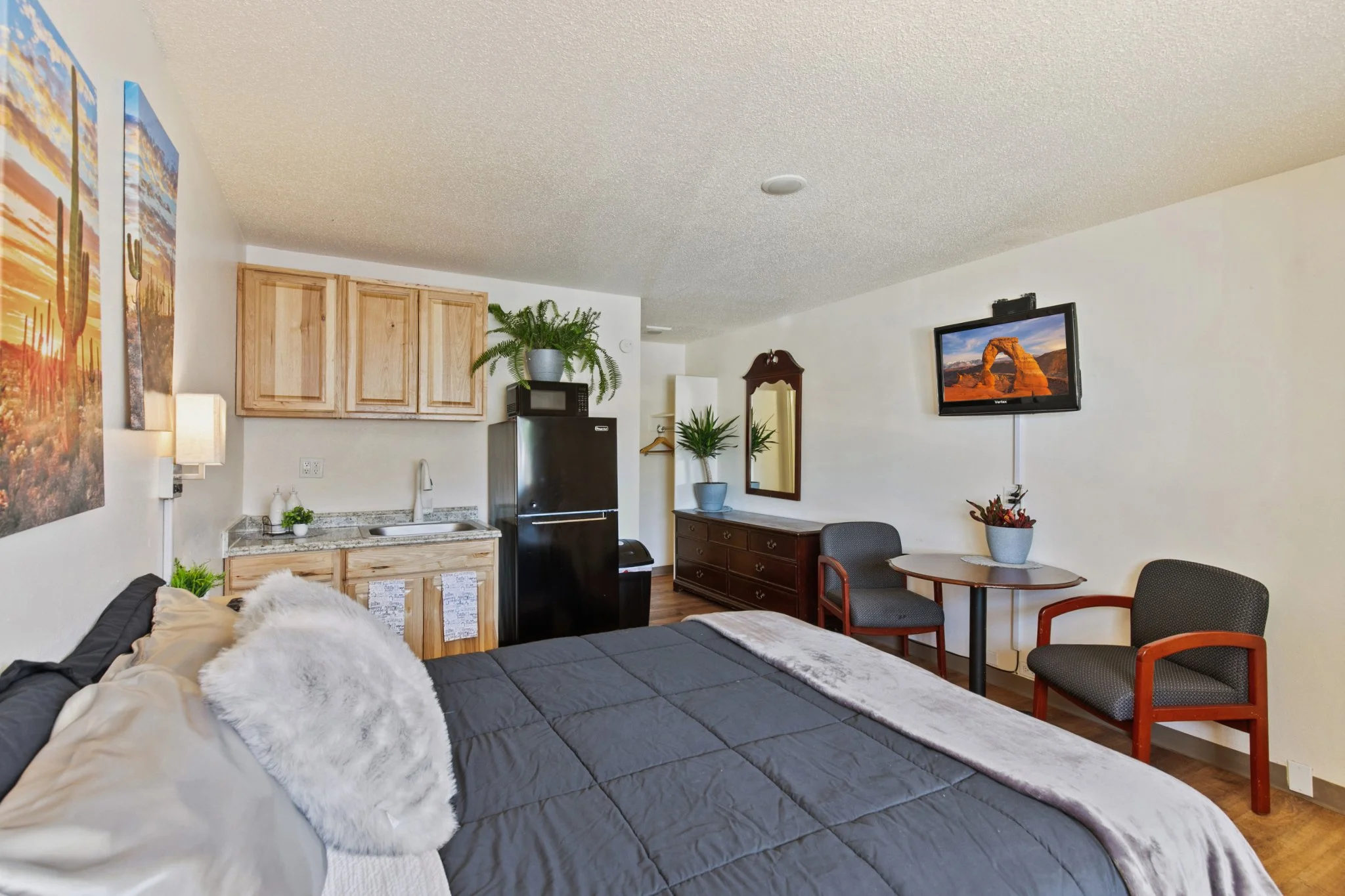 Studio apartment with a bed in the foreground, a small kitchenette with cabinets, a refrigerator, a microwave, and a potted fern, a wall-mounted TV, a mirror, a dining table with chairs, and wall art depicting a desert scene.