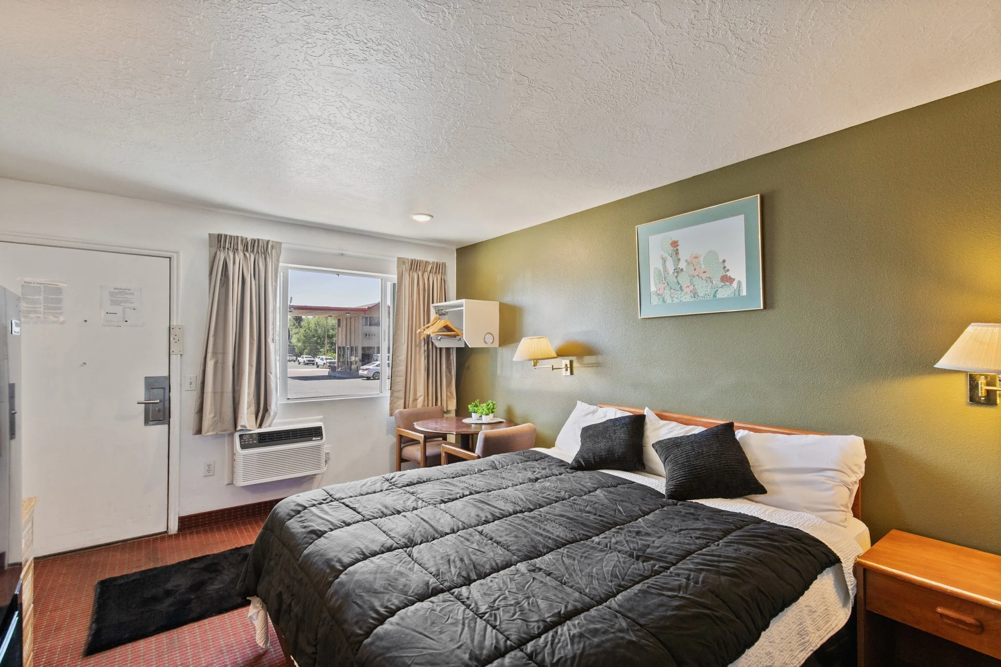 A hotel room with two beds, a small table with two chairs, a window with beige curtains, a wall mirror, and framed wall art with cactus designs.