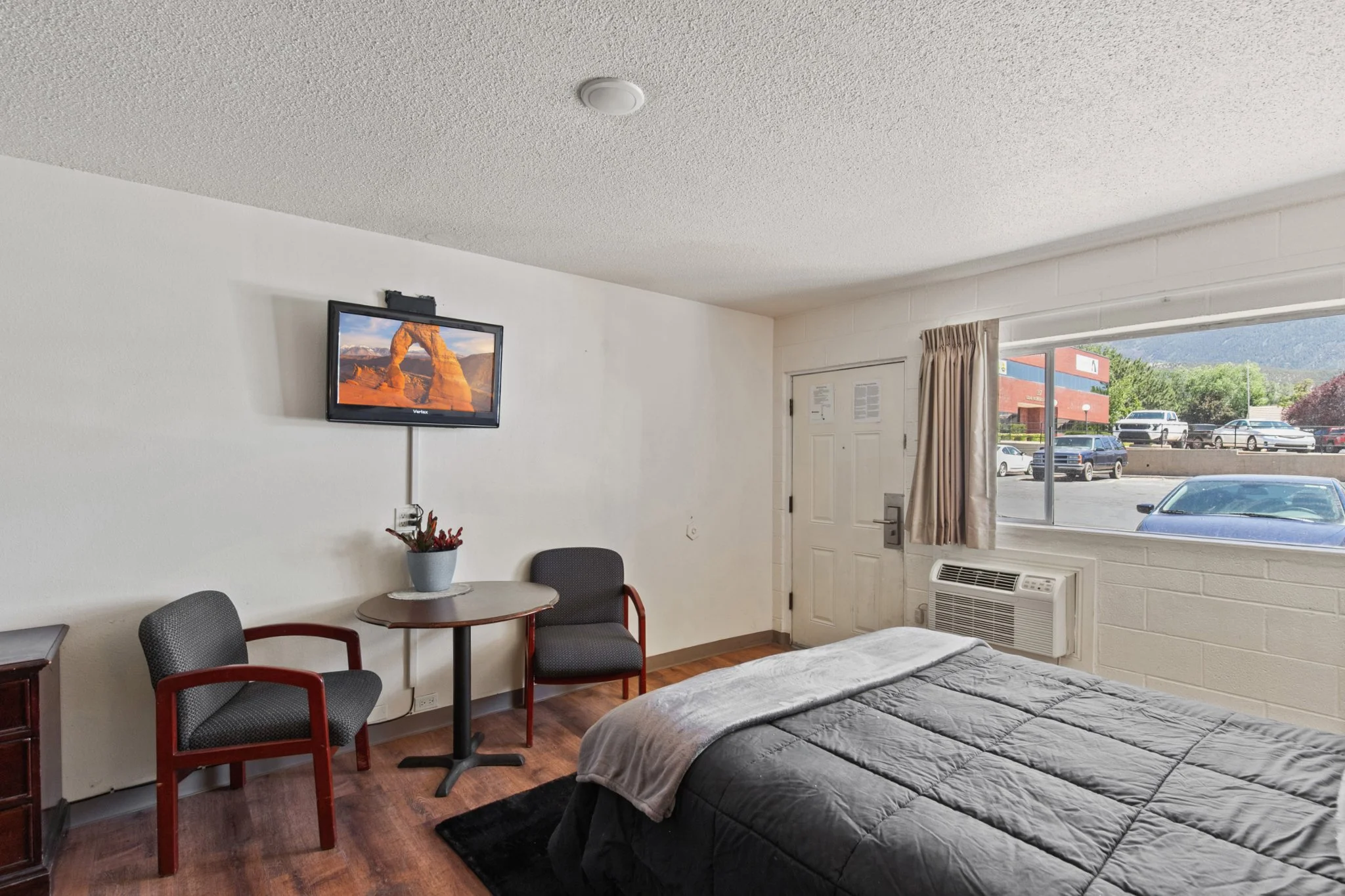 A room with a bed, a small round table with two chairs, a wall-mounted TV showing a desert landscape, a window with curtains, and cars parked outside visible through the window.