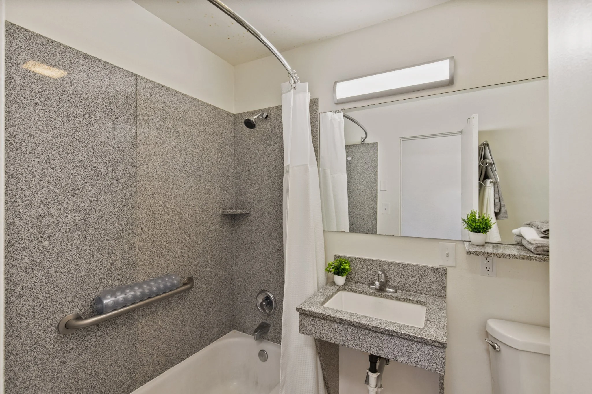 A bathroom with a walk-in tile shower, a granite vanity with a sink, a mirror, a potted plant, and towels on a shelf.