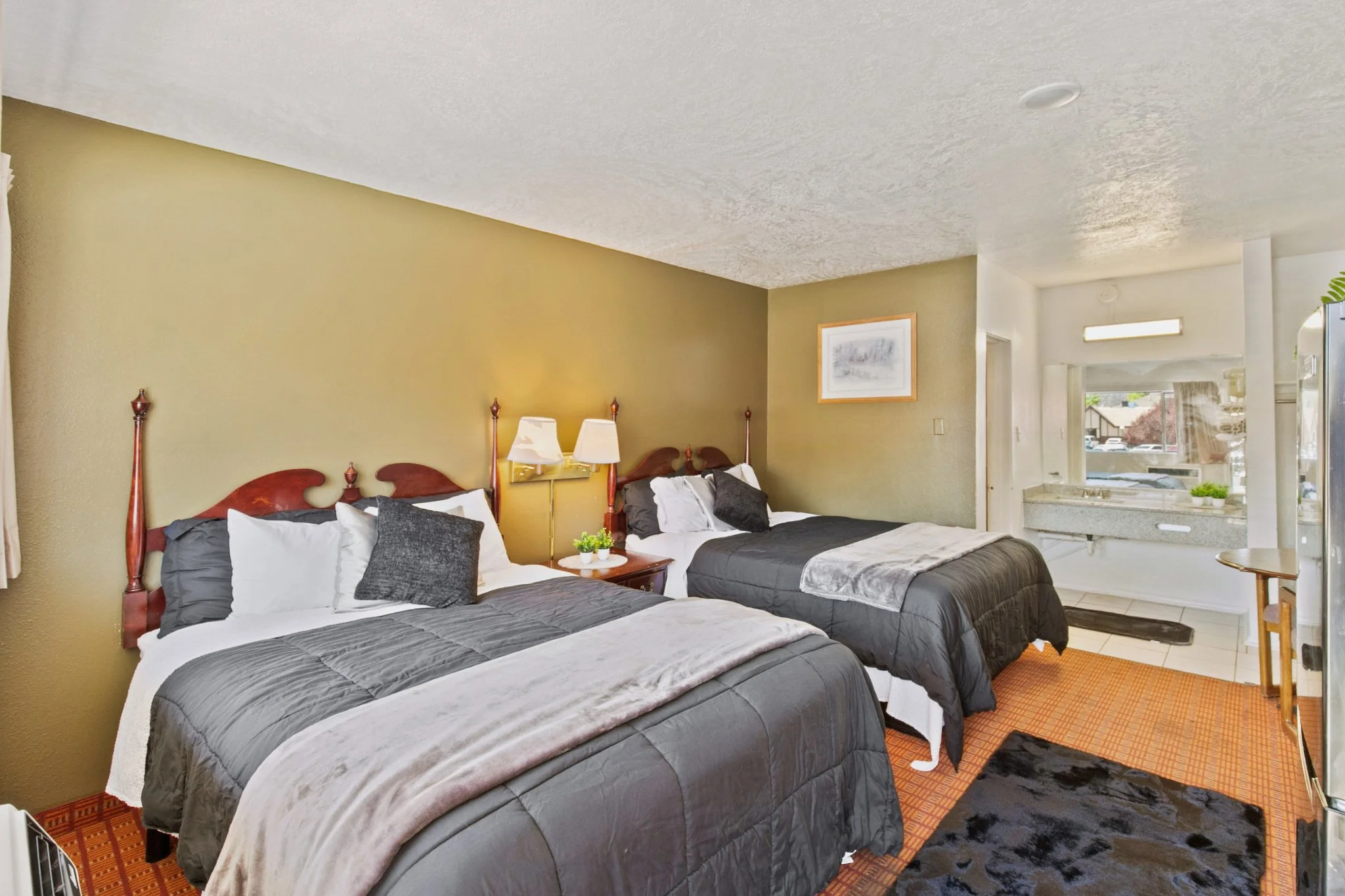 A hotel room with two double beds, each with white and dark gray bedding and pillows, a wooden headboard, a wall-mounted lamps, and a framed picture on a yellow wall. In the background, a kitchenette with a sink, countertop, and window with curtains 