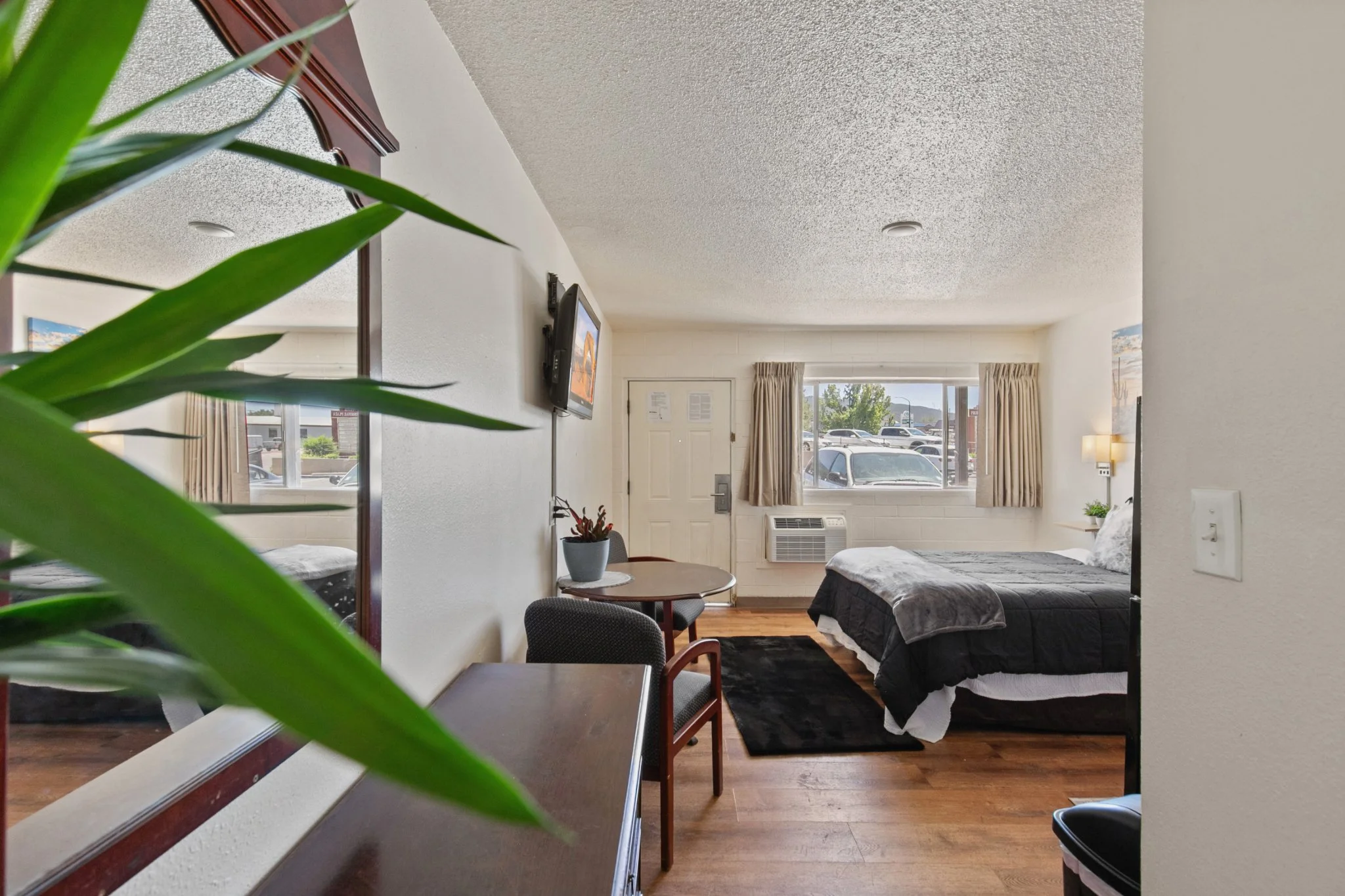 A view of a hotel room with a bed, a small table, chairs, and a window with curtains. Part of a green plant and a mirror frame are visible in the foreground.