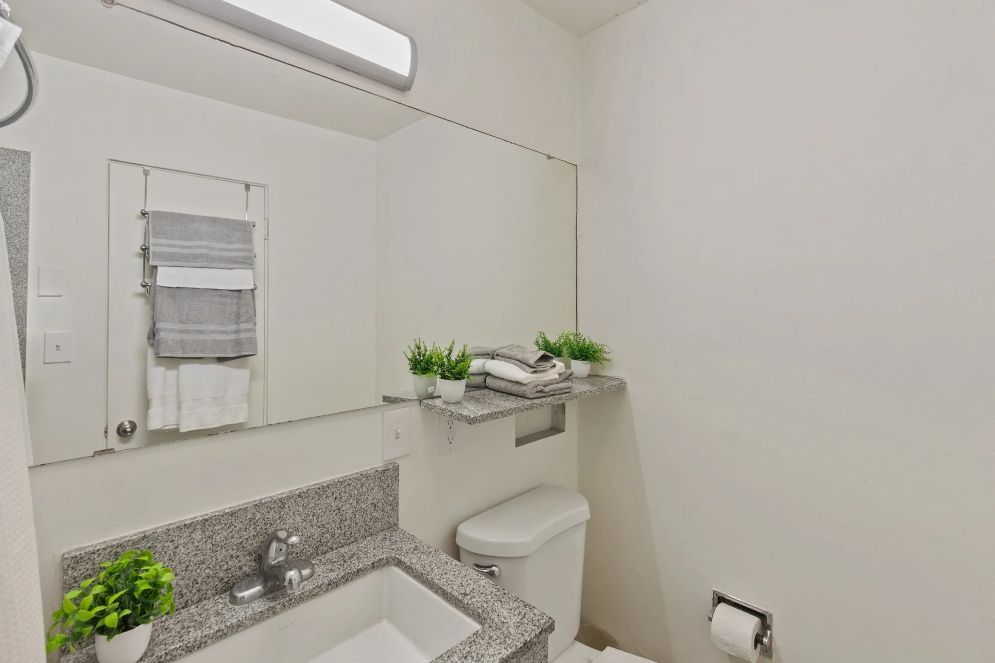 A small bathroom with a granite sink countertop, a white toilet, a mirror with a towel rack, small potted plants, and some neatly folded towels on a granite shelf.