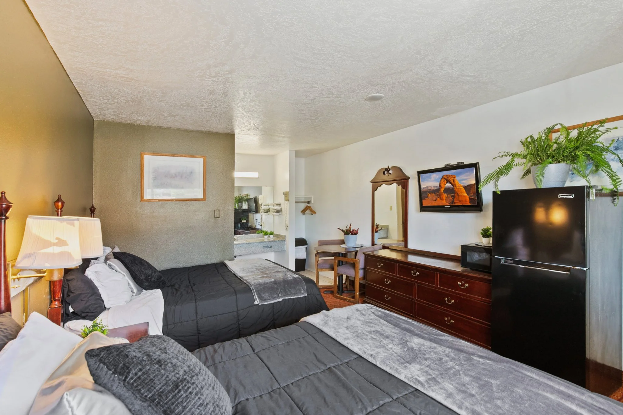 Hotel room with two beds, a wooden dresser with a TV on top, a mirror, and a large potted plant. The room has a small dining table, chairs, and a wall with framed pictures.