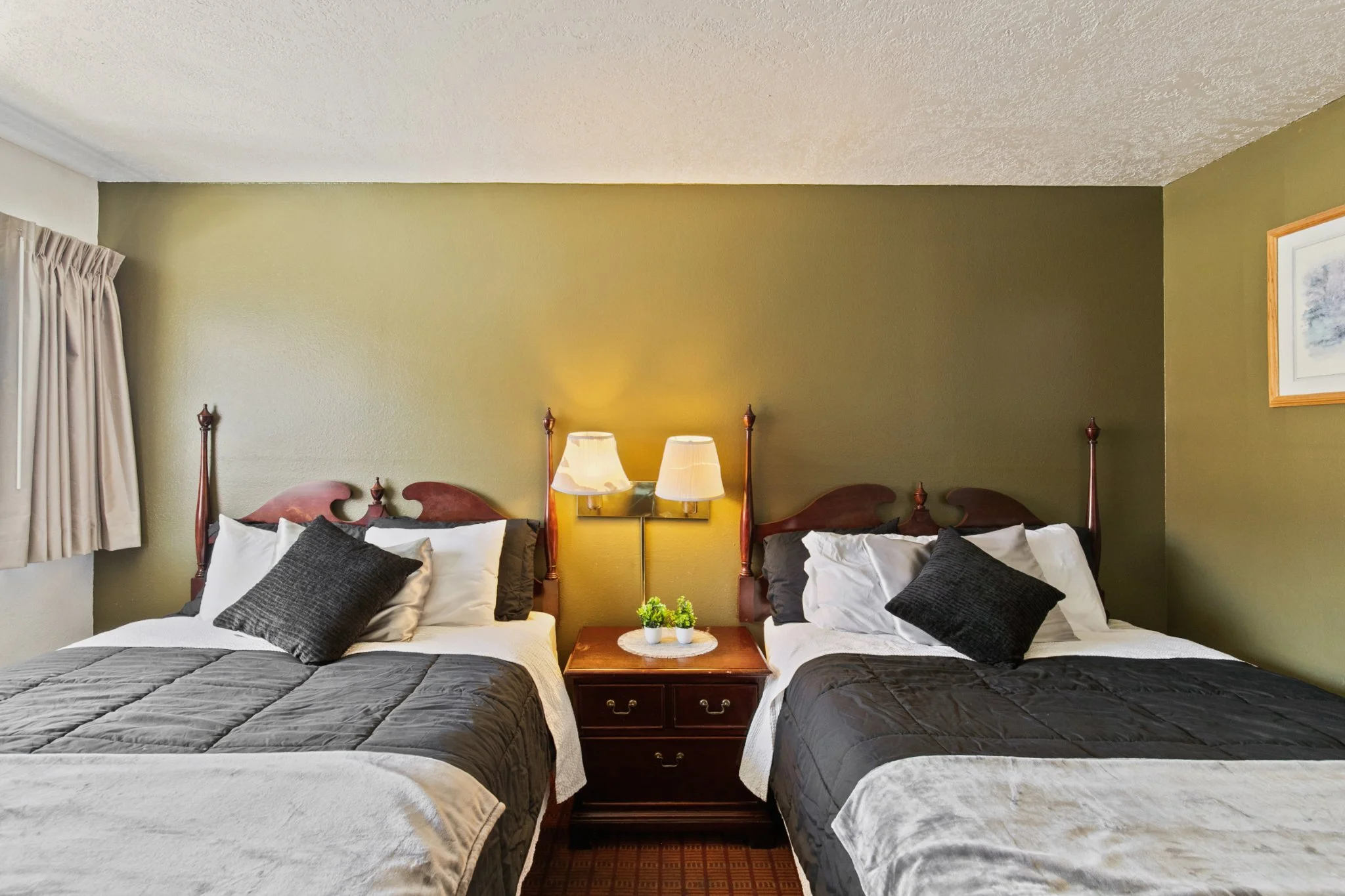 Two double beds in a bedroom with a shared wooden nightstand, two lamps, pillows, and a small plant, with a wall-mounted picture and curtains on the left side.