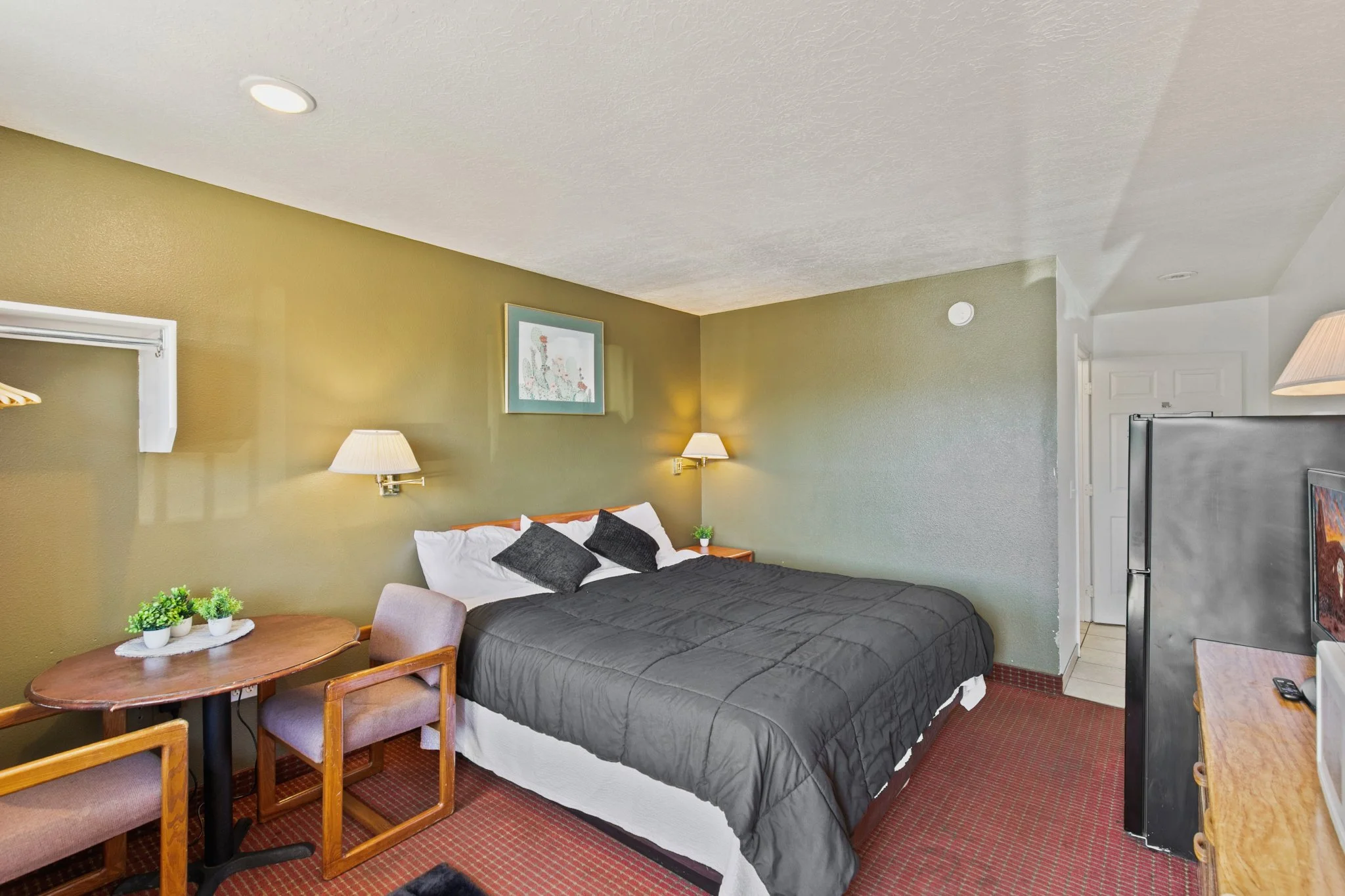 A hotel room with a bed, a table with chairs, a wall-mounted lamp, and a refrigerator. The room has green walls, a framed artwork, and a corner with a door.
