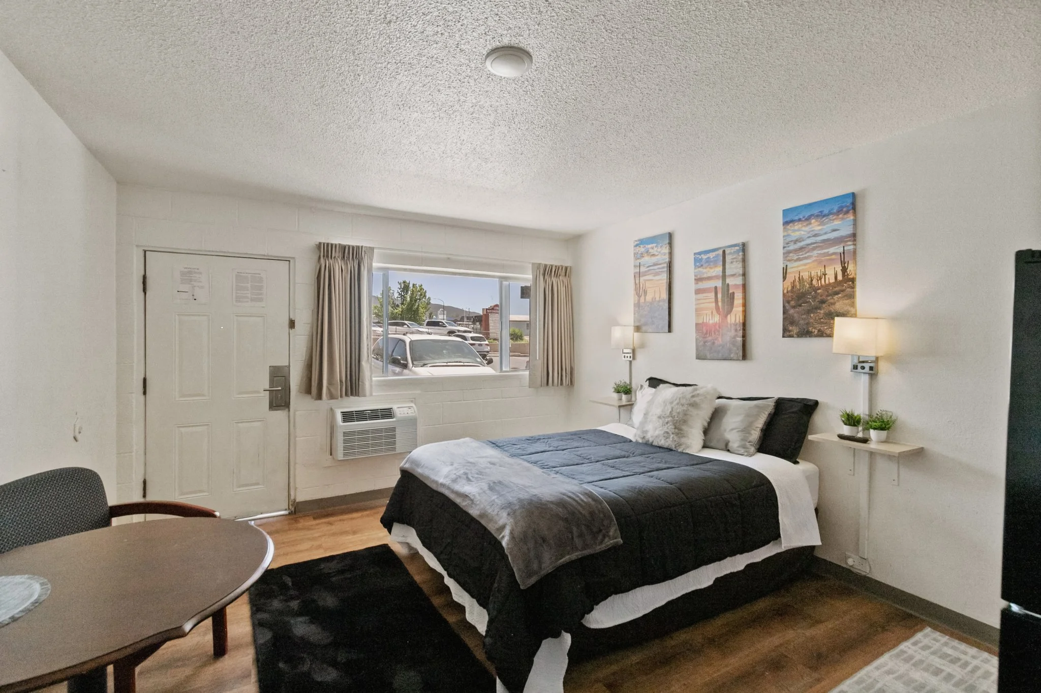 Interior of a studio apartment with a bed, small side tables with potted plants, wall art, a window with curtains, and a door.