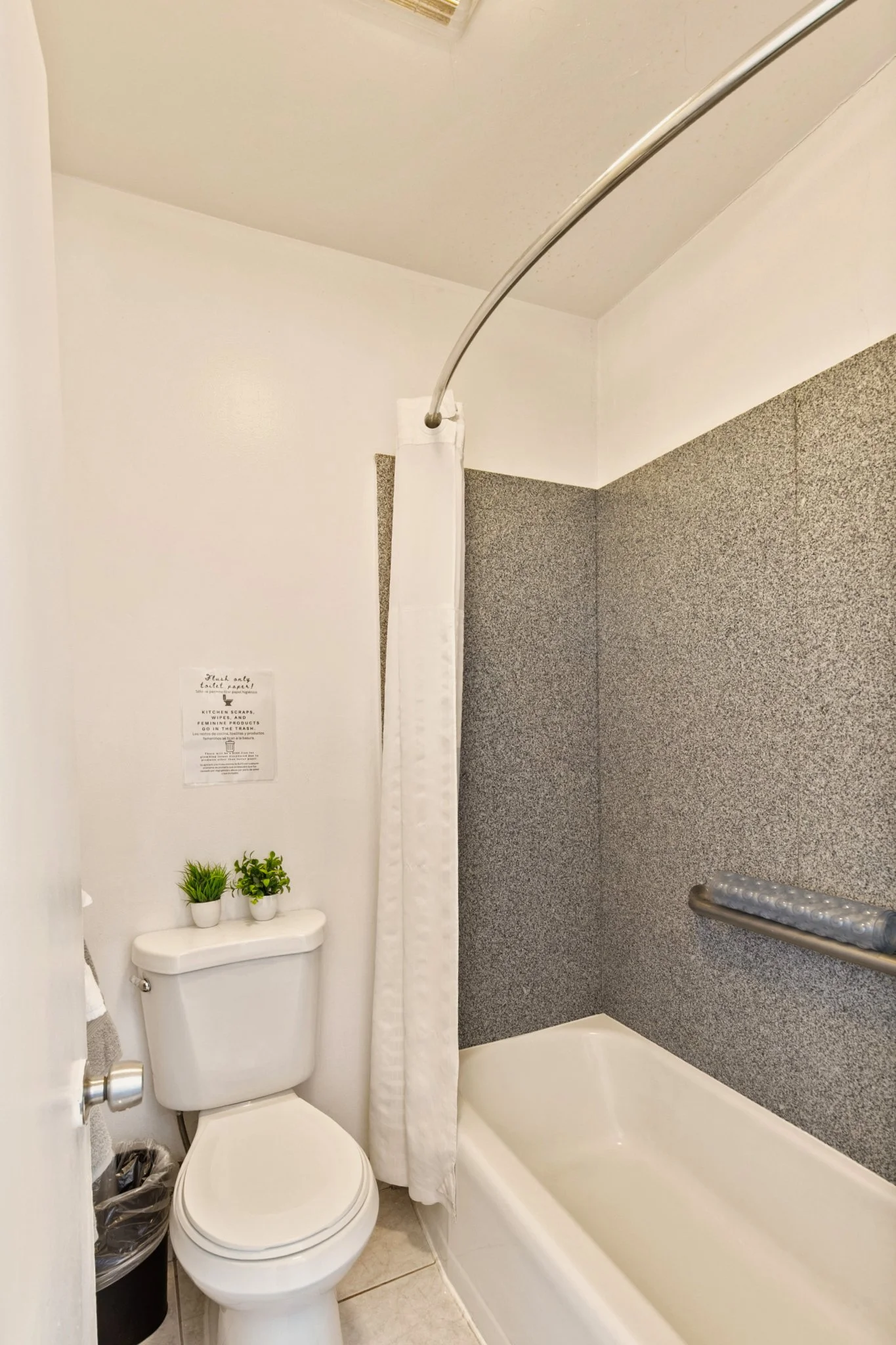 A small bathroom with a toilet, bathtub, and a shower curtain. Two small potted plants on top of the toilet tank and a gray textured wall. A towel hanging on a bar, a trash can beside the toilet, and a sign on the wall.