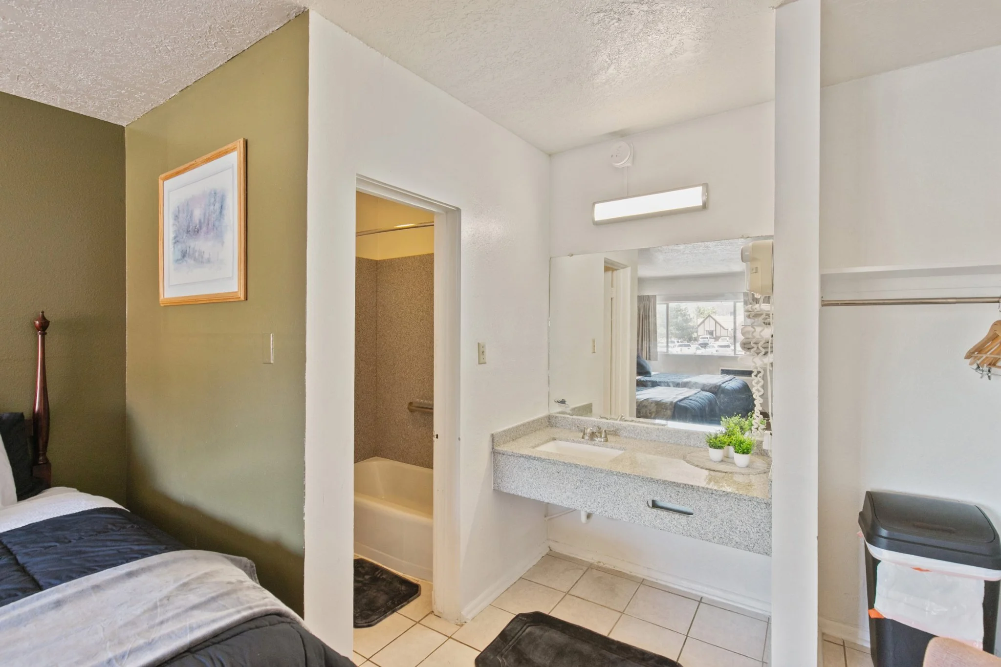 Interior view of a motel room with a partial bed, a mini kitchen area with a sink, small plants, and a mirror with a view of a parking lot and building outside.