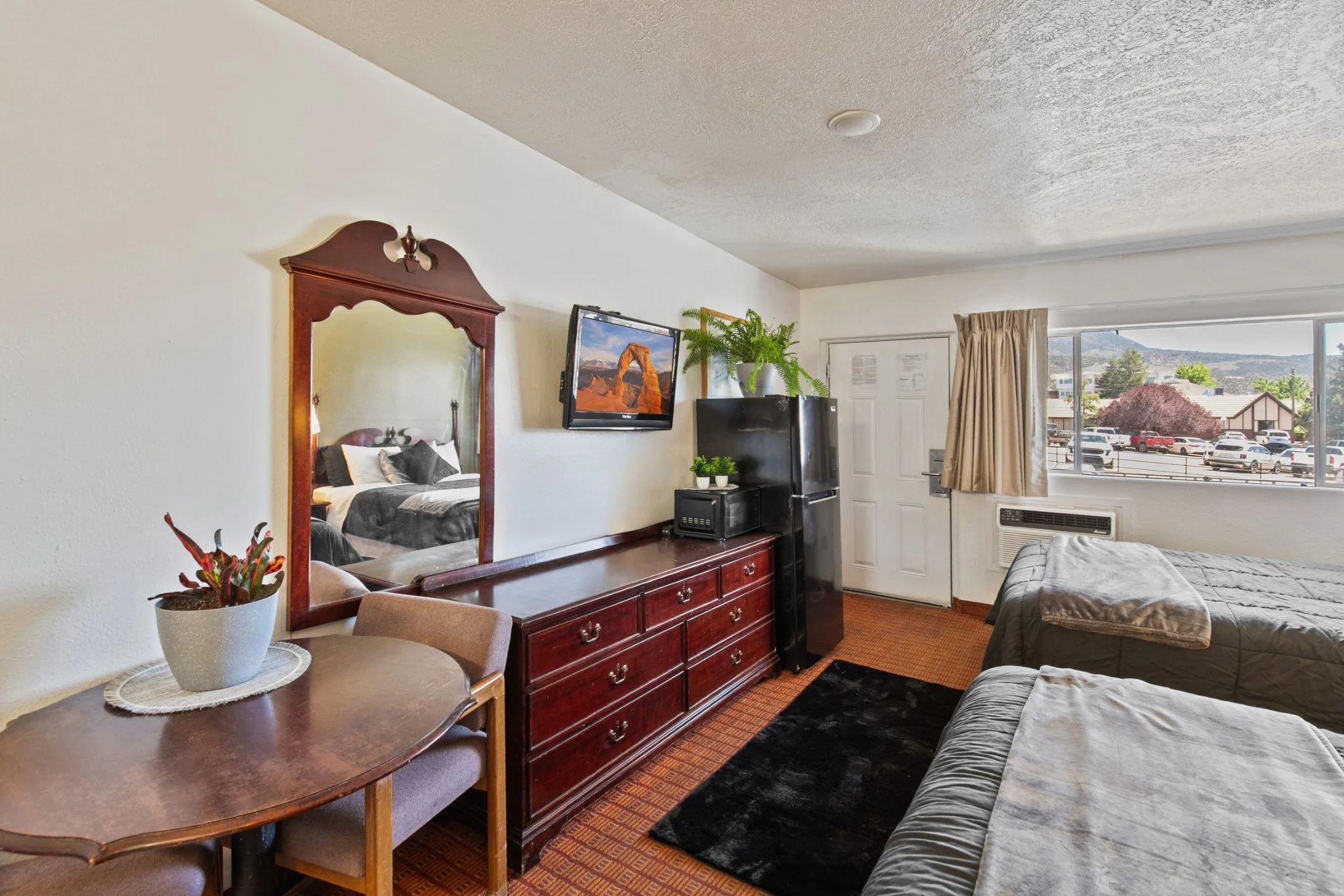 Inside a hotel room with a bed, a dresser with a mirror, a small table with a potted plant, a television on the wall, and a window showing a parking lot and trees outside.