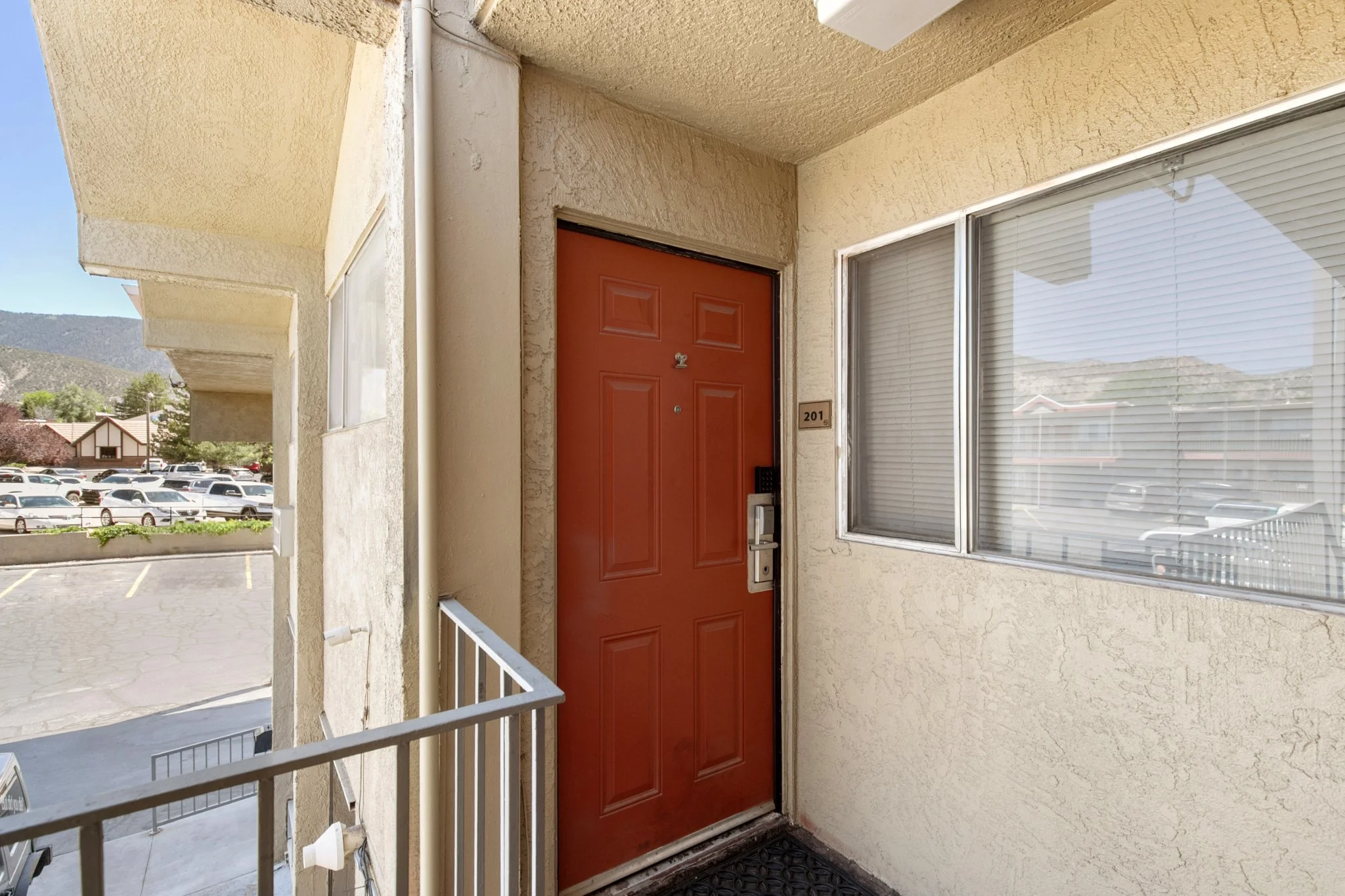 The image shows the front entrance of an apartment or condo unit with a bright orange door, a small number plate reading '201', and a window with closed blinds beside the door. The building's exterior walls are beige with a textured finish. There is 