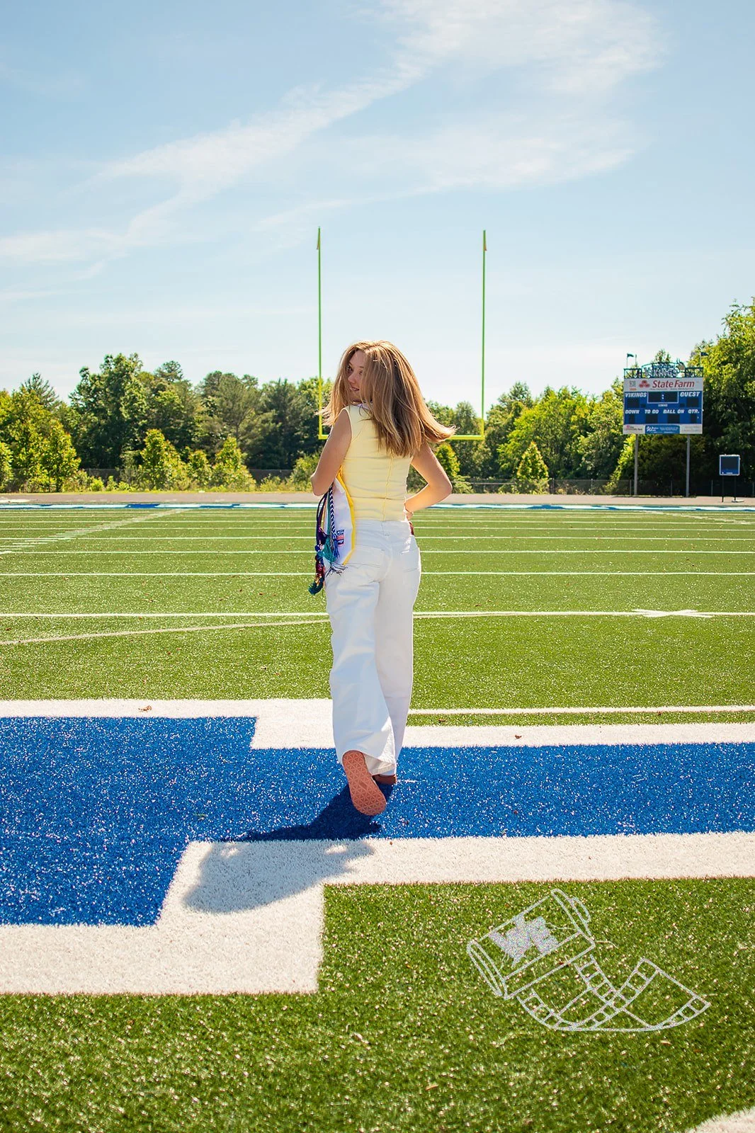 A young woman with long hair, wearing a yellow sleeveless top and white pants, standing on a football field with a scoreboard and goalposts in the background. She is looking back over her shoulder.