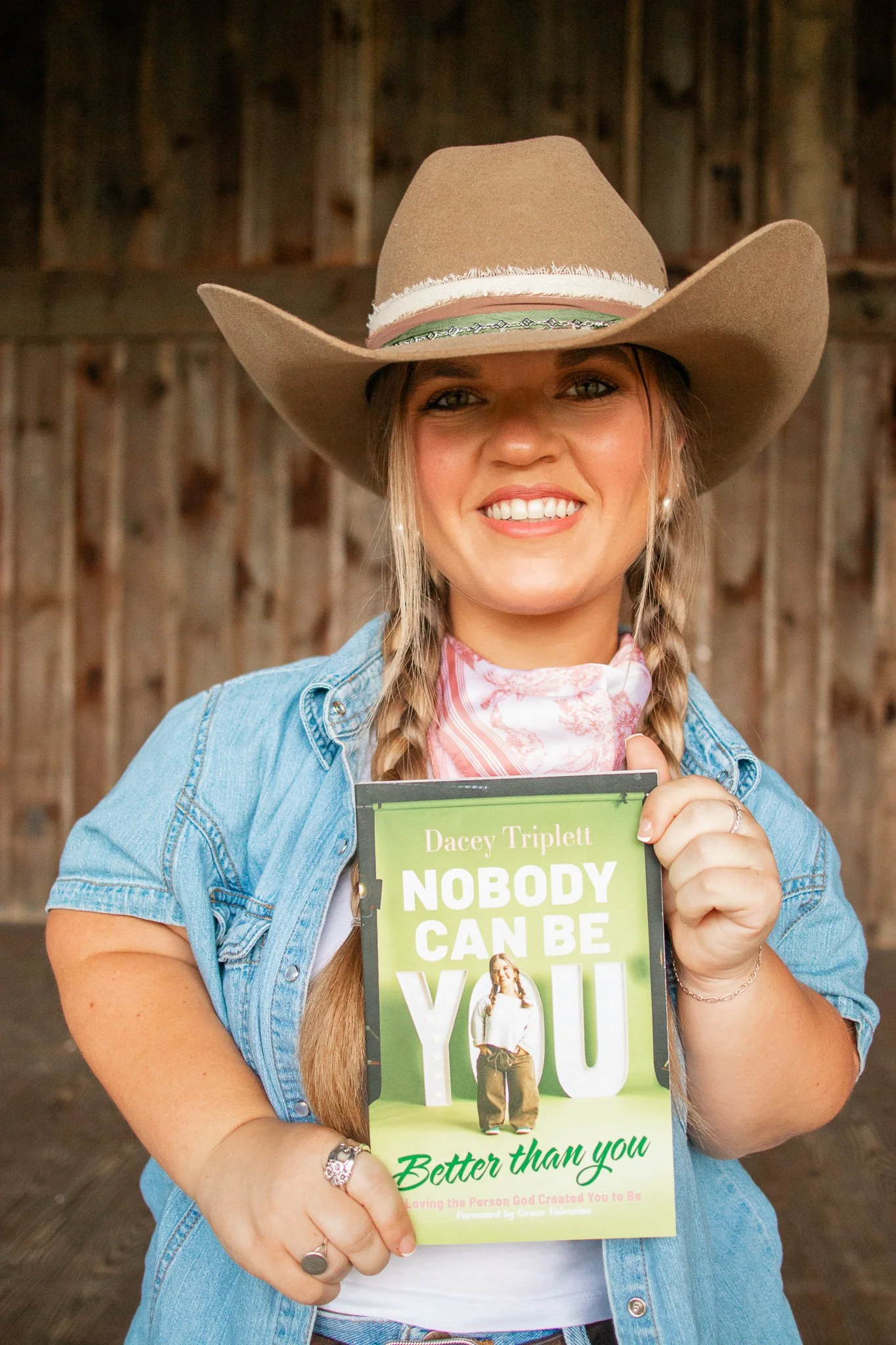 A smiling woman with braided hair wearing a cowboy hat and denim jacket holding a book titled 'Nobody Can Be You' in front of a wooden background.