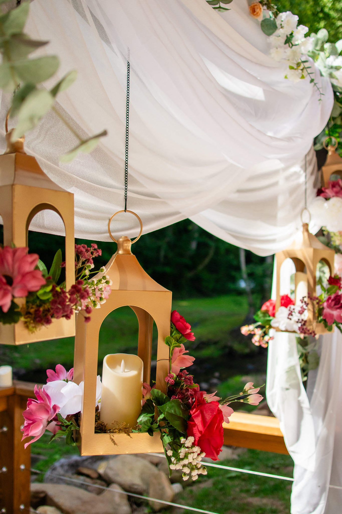 Decorative hanging lanterns and floral arrangements with pink, red, and white flowers, draped white fabric, and candles, likely part of an outdoor wedding or event setup.