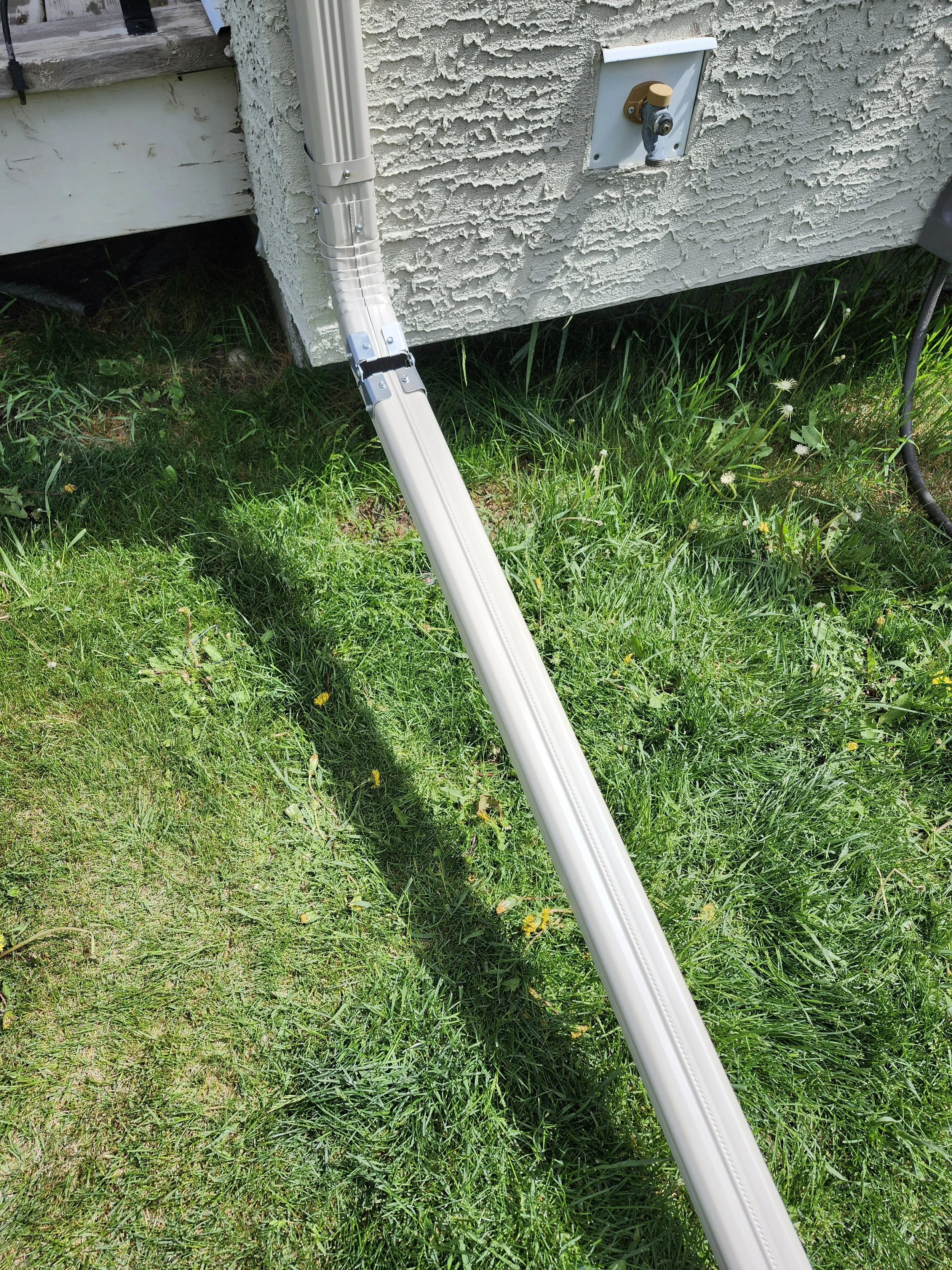 A white, retractable, metal multi-angle ladder leaning against a house with weathered grayish siding and a small patch of grass with dandelions and other plants.
