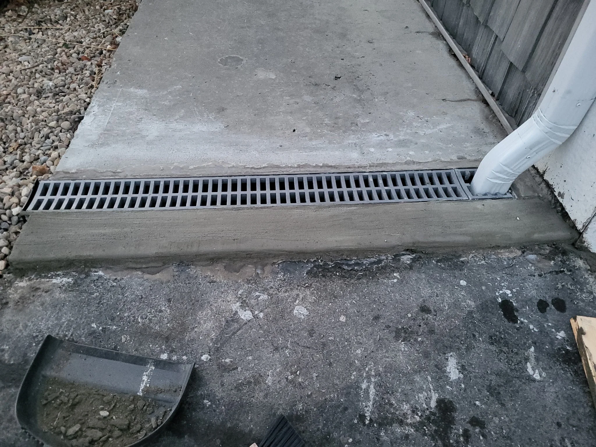 Concrete sidewalk with drainage grate, white plastic pipe, and gravel area on the left side, and a wooden fence on the right side.