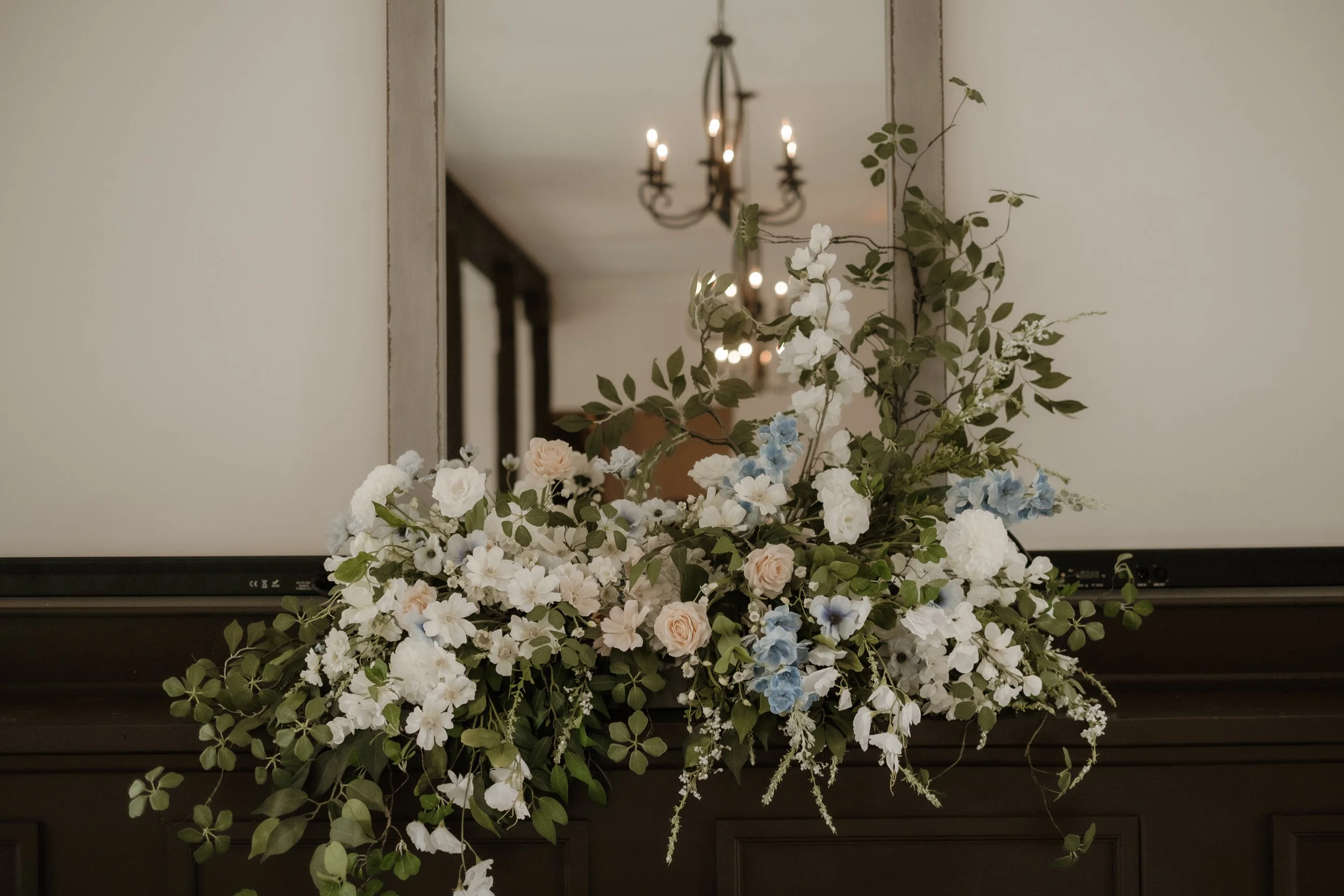 A bouquet of white and pale pink roses, blue and white flowers, and green leaves resting on a black surface in front of a mirror in a room with a chandelier.
