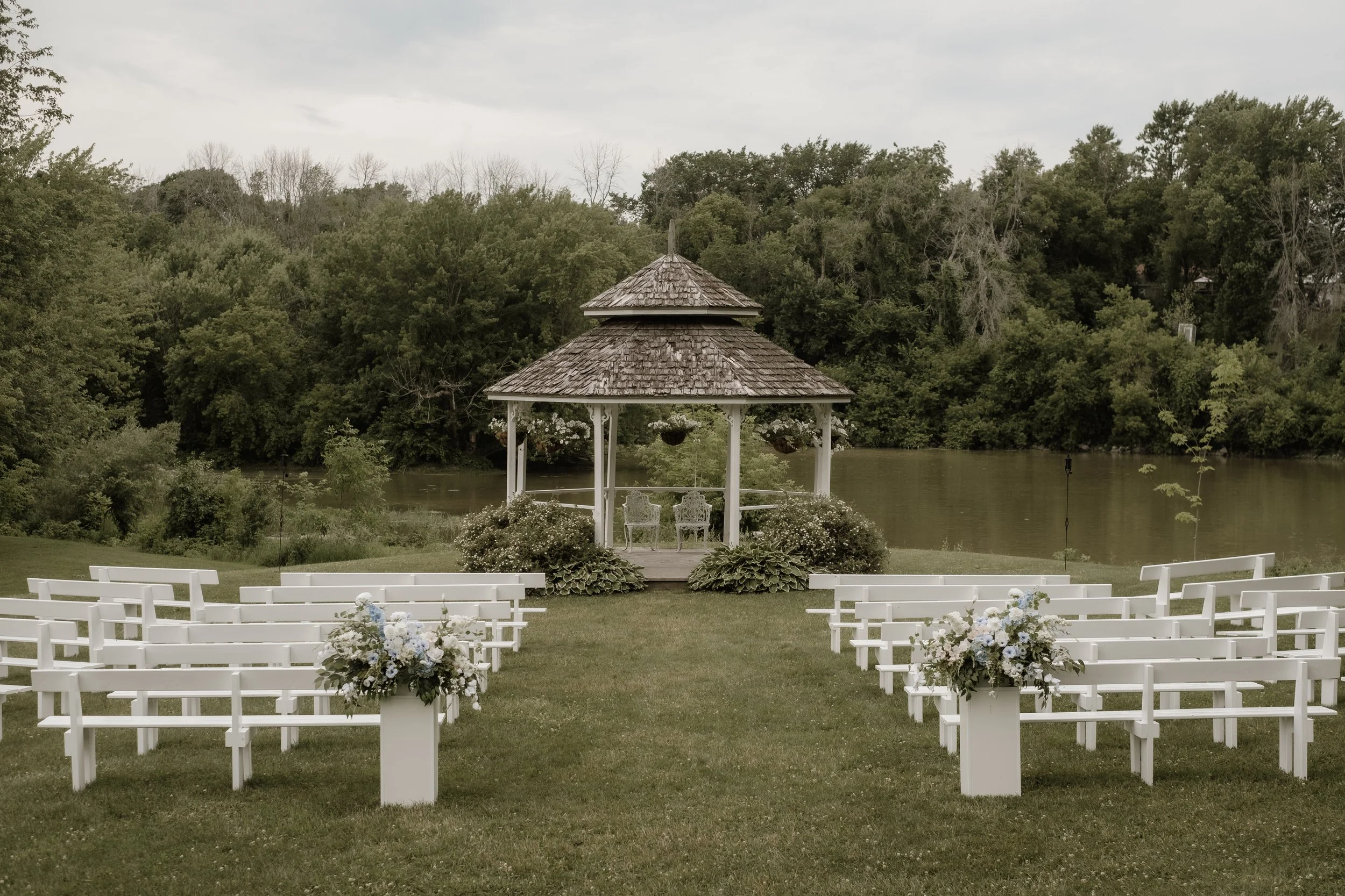 Outdoor wedding ceremony setup with white chairs, flower arrangements, a gazebo, a pond, and lush trees in the background.