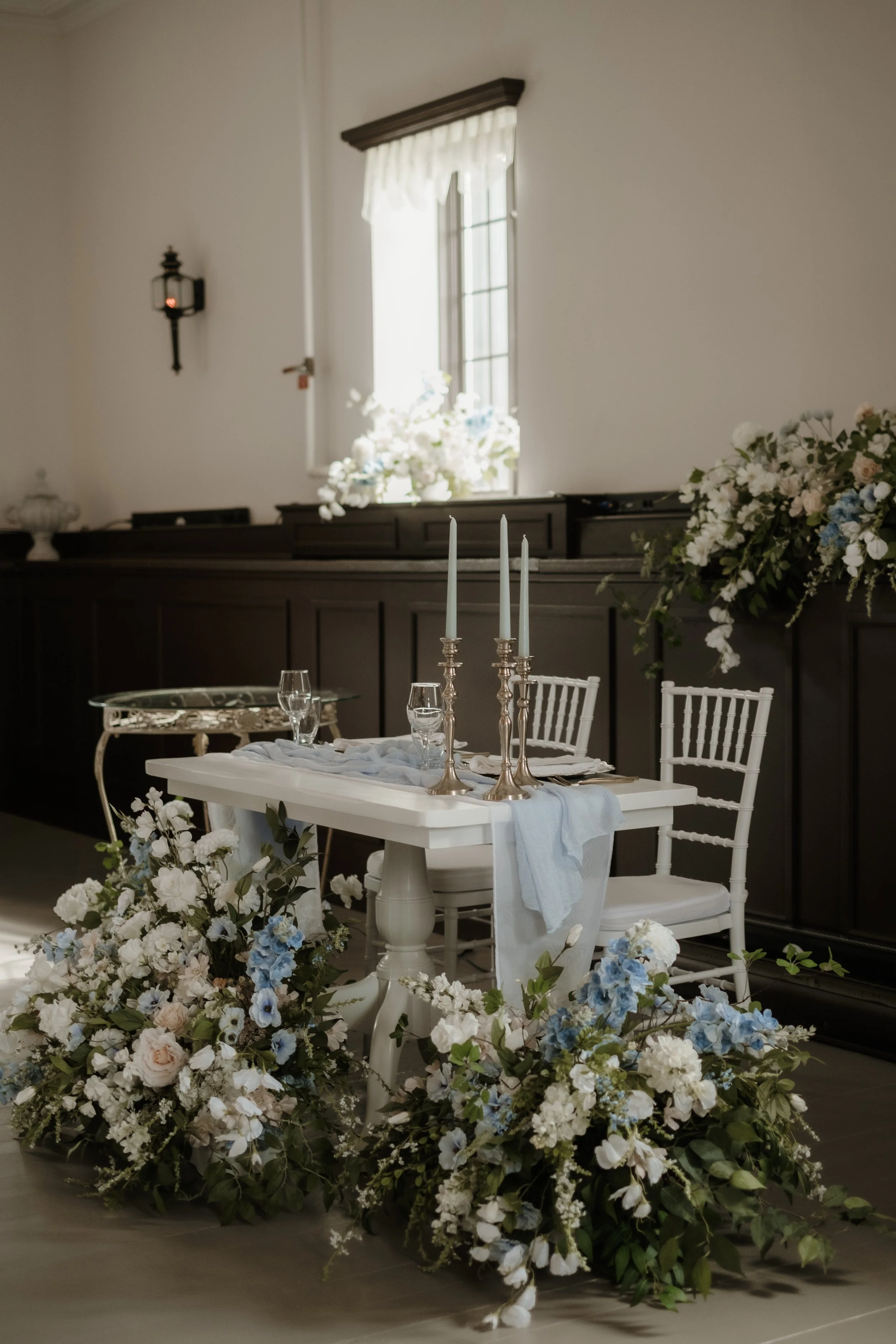 Elegant table setting with white chairs, tall candlesticks, glassware, and floral arrangements in white, blue, and blush colors, set in a room with dark wood paneling and a window with a white valance.