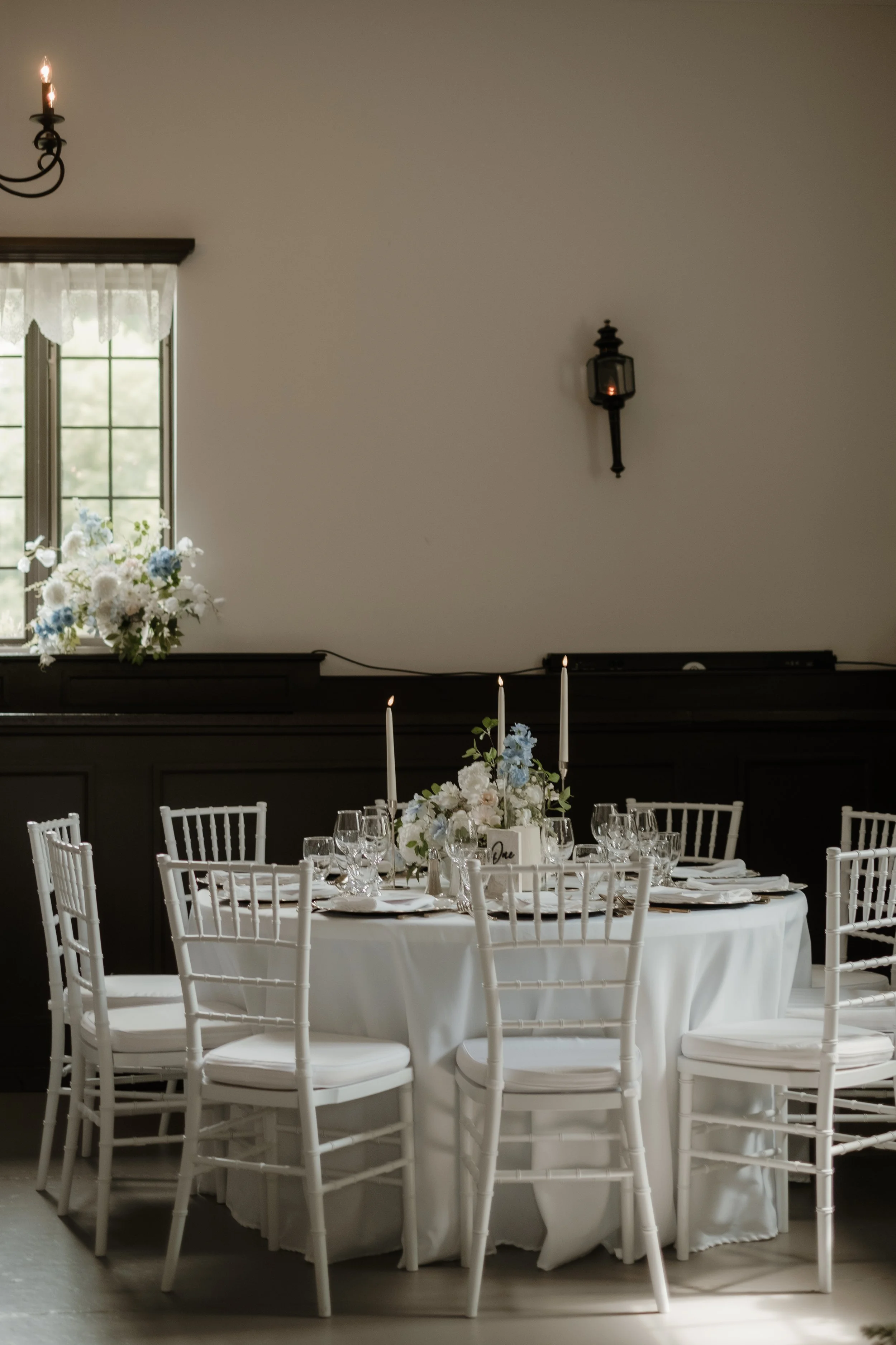 Elegant dining room with a round table set with white tablecloth, glassware, napkins, and a floral centerpiece, surrounded by white chairs, with a window and floral arrangement in the background.