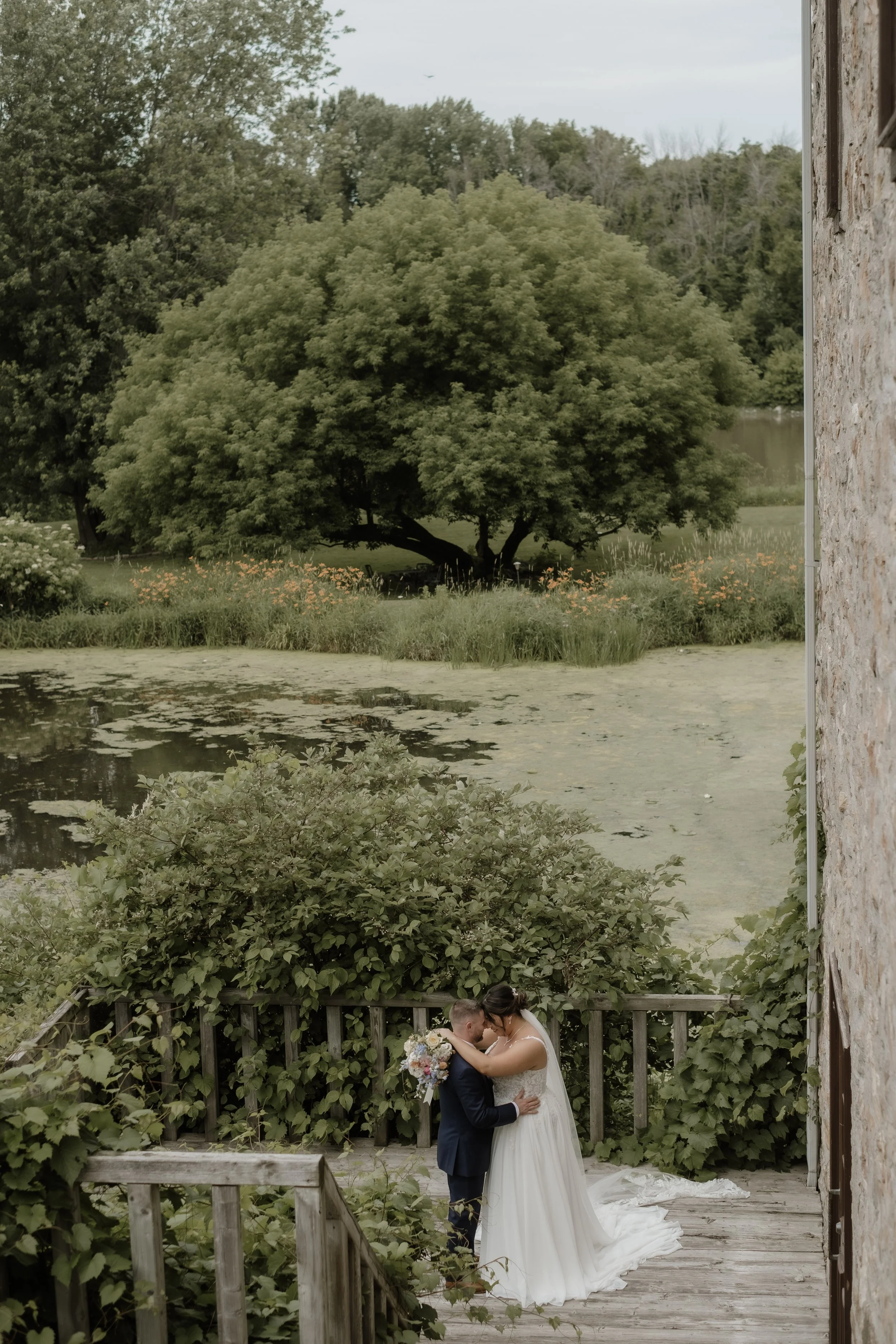 A bride and groom embrace on a wooden deck outdoors, with lush greenery, trees, a pond, and cloudy sky in the background.