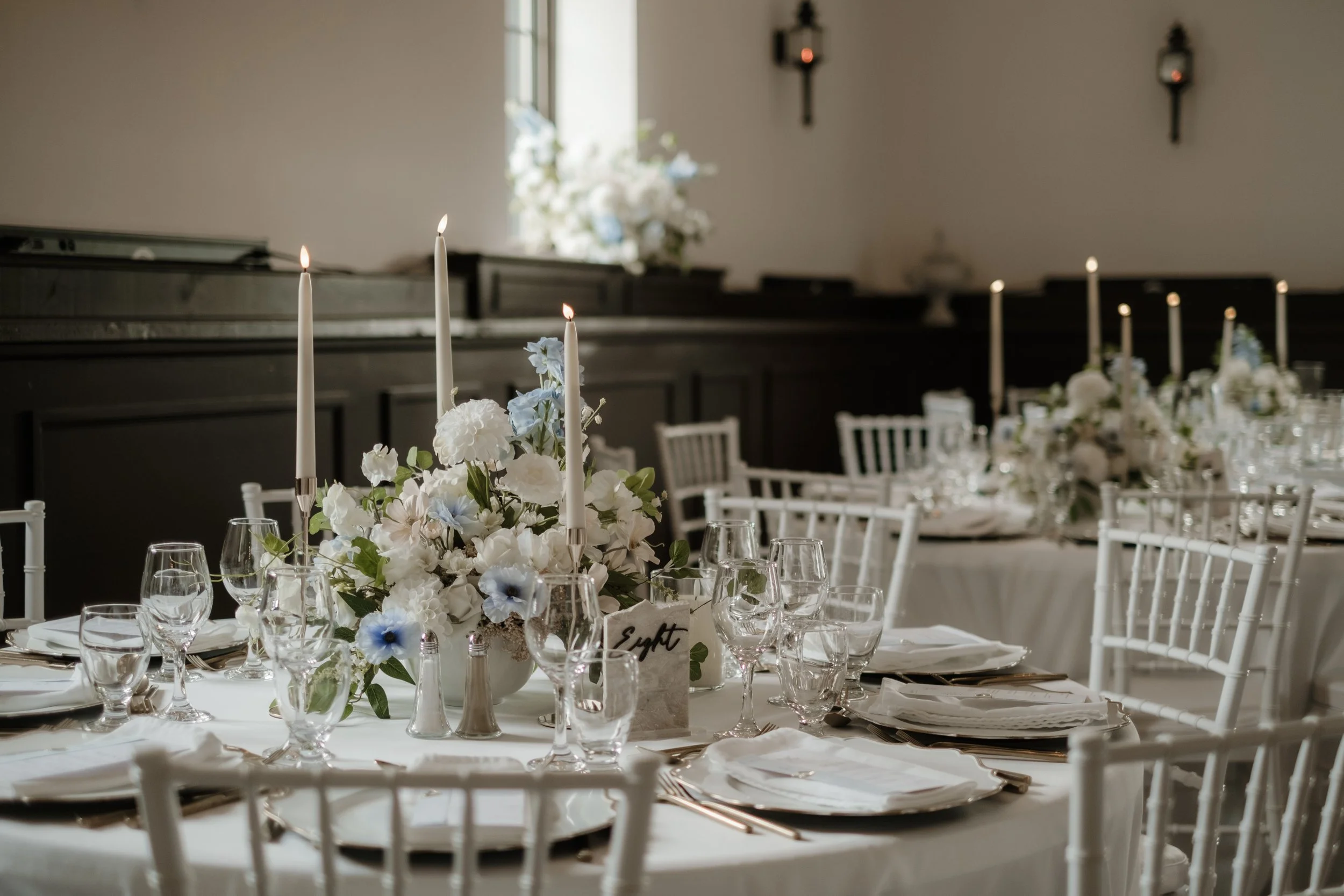 Elegant banquet table with a floral centerpiece, tall white candles, glassware, and white chairs in a decorated room with candle sconces on the wall.
