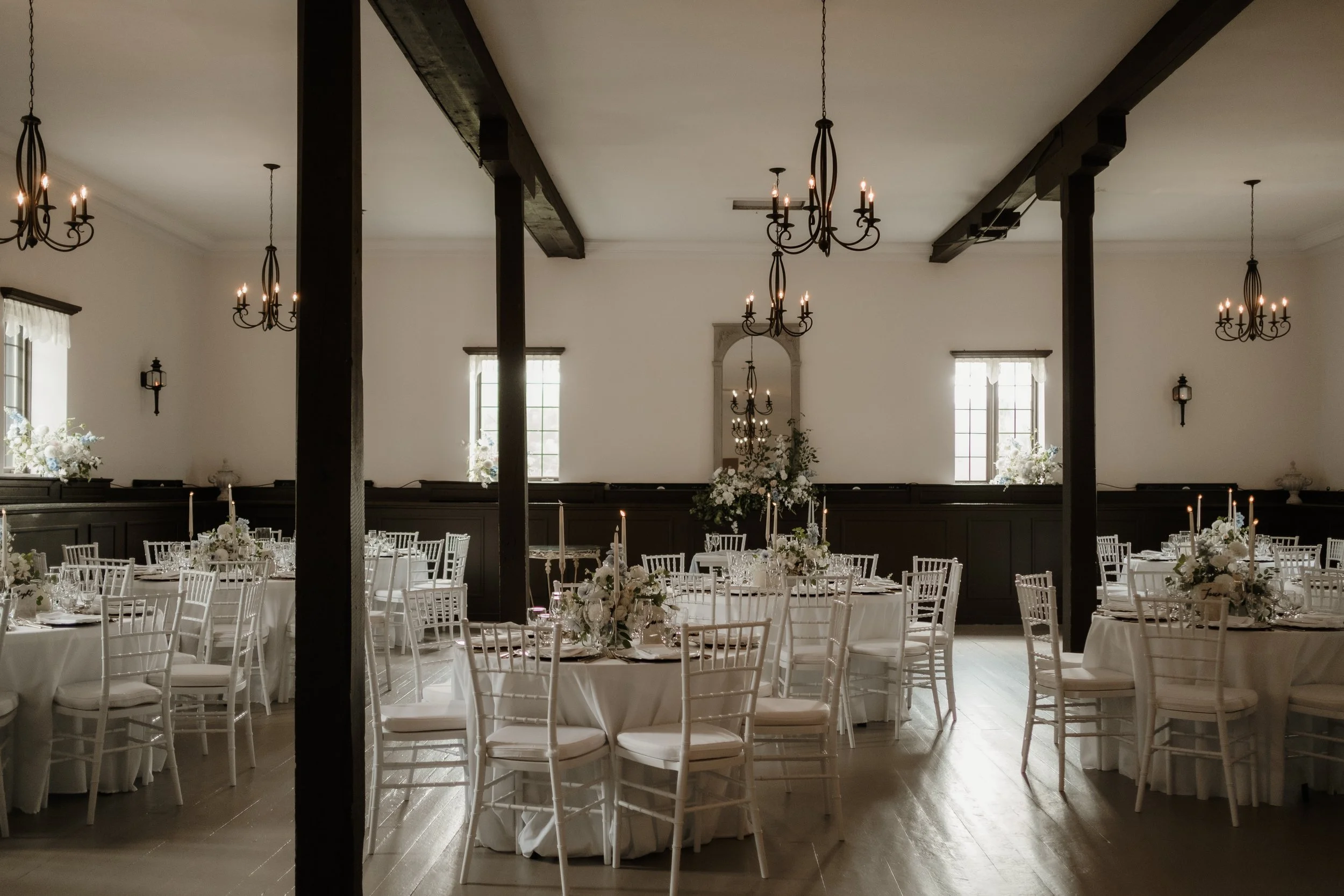 Elegant banquet hall set up with round tables, white chairs, floral centerpieces, and chandeliers hanging from the ceiling.