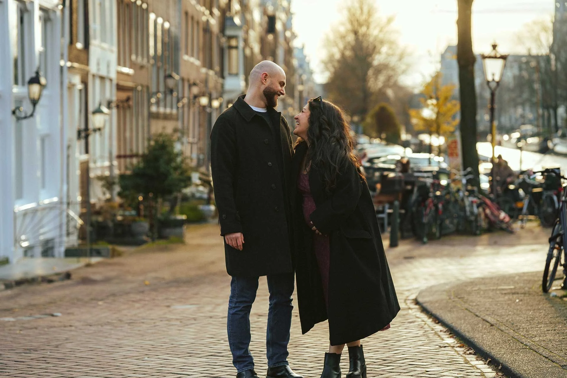 A full-length shot of a pregnant woman and a man standing on a cobblestone street in an urban setting, looking lovingly at each other. The woman is wearing a long dark coat and the man is wearing a dark jacket