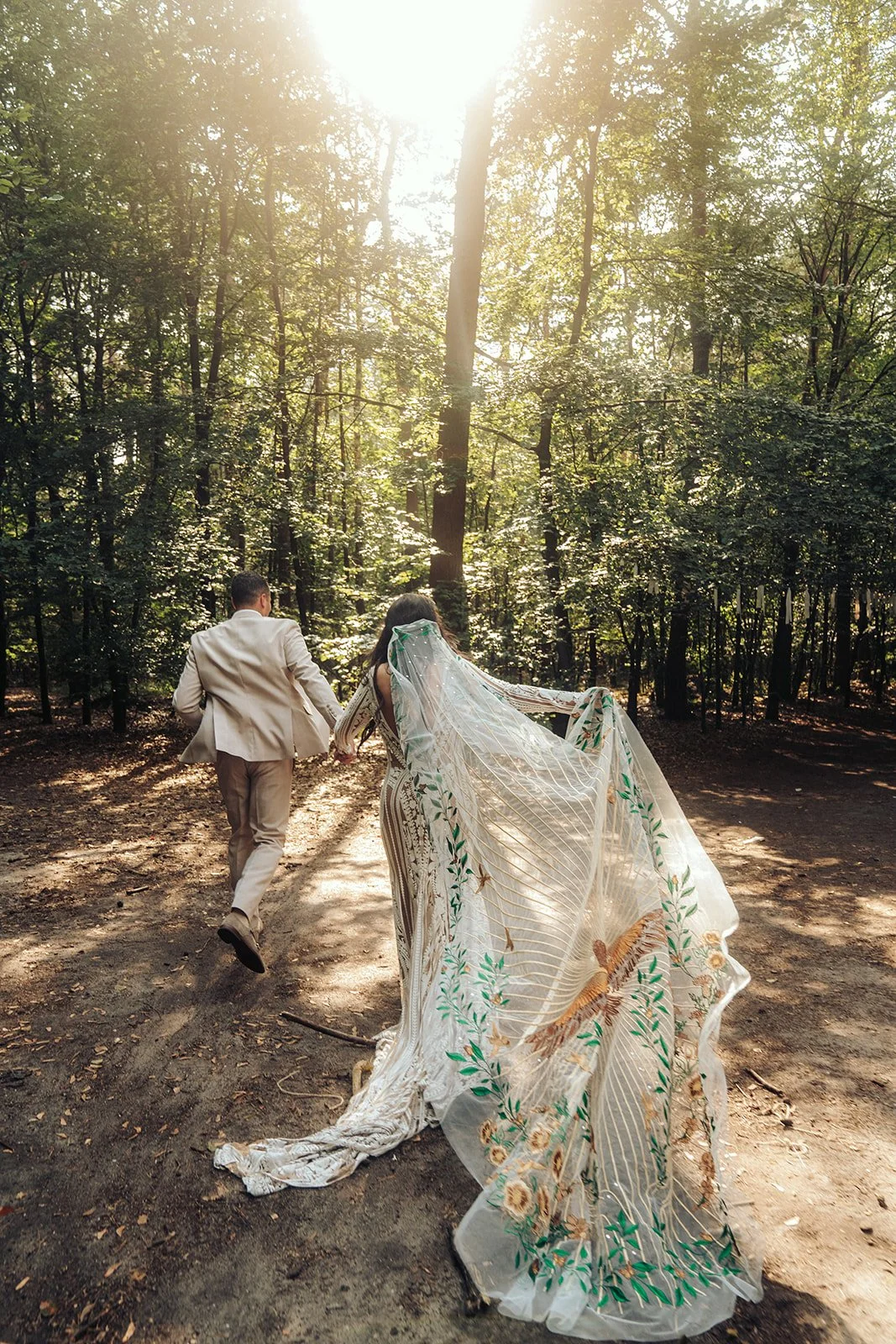 Golden light filters through tall trees as a couple, deeply in love, runs through a forest path, their unique bridal attire flowing – a moment of natural wedding beauty in the Dutch countryside.