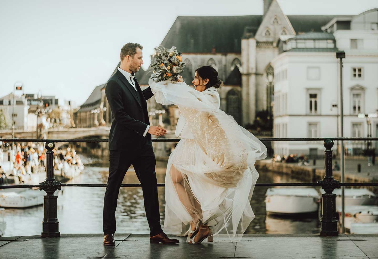 A playful moment between a bride and groom on a bridge over a canal. The bride, in a white dress, holds a bouquet while the wind blows her dress. The groom, in a black suit, stands facing her.