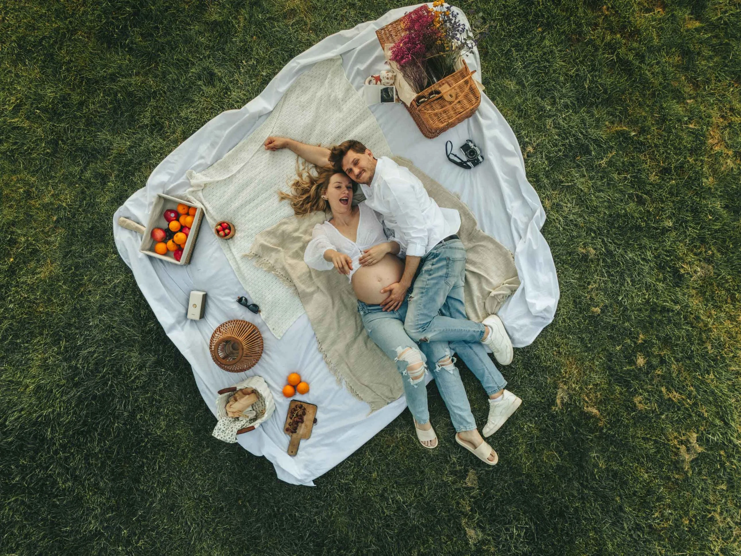 An aerial view of a pregnant couple lying on a white picnic blanket on green grass. They are cuddling, surrounded by fruit, pastries, and a picnic basket.