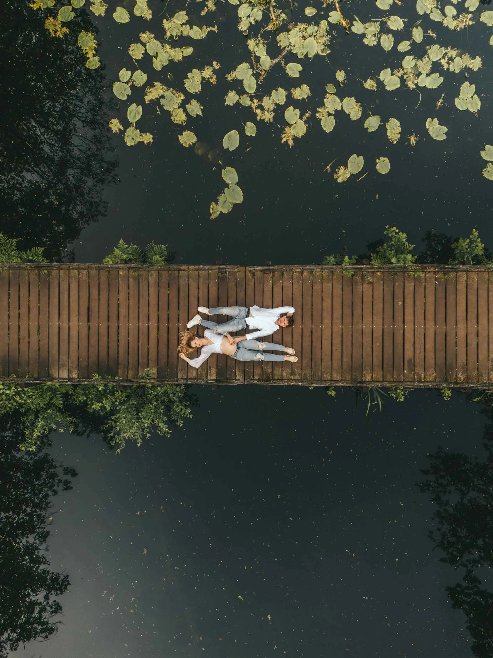 An aerial view of a pregnant couple lying on a wooden bridge over dark water with lily pads. They are looking up, dressed in light colored clothes.