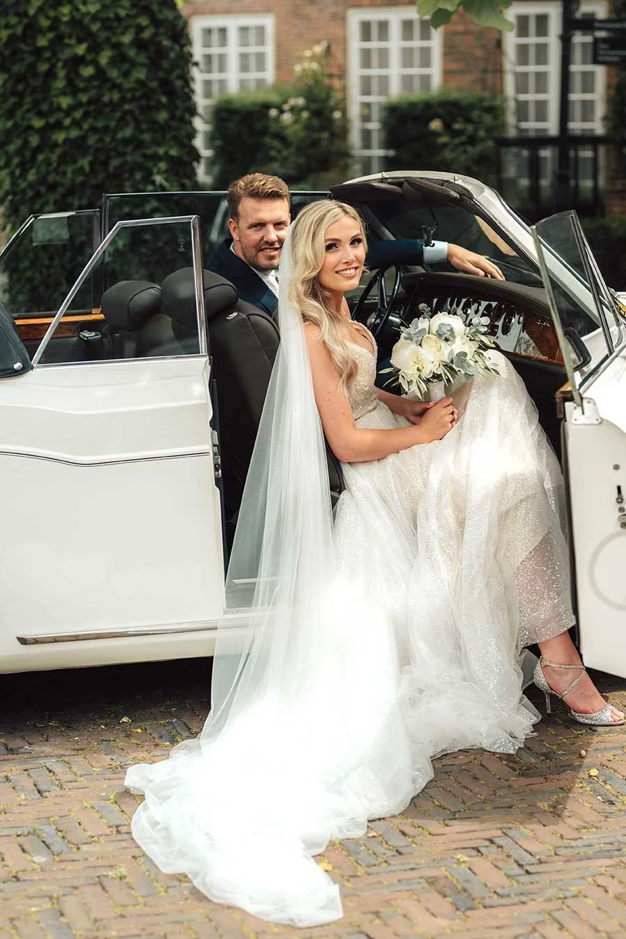 A smiling bride in a sparkling white wedding dress with a long veil sits in a classic white convertible car, holding a white bouquet, while the groom smiles from the driver's seat.